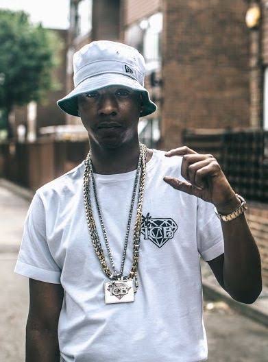 Nines wearing a white bucket hat and white t-shirt, making a gesture with his left hand, photographed outdoors against a brick background.