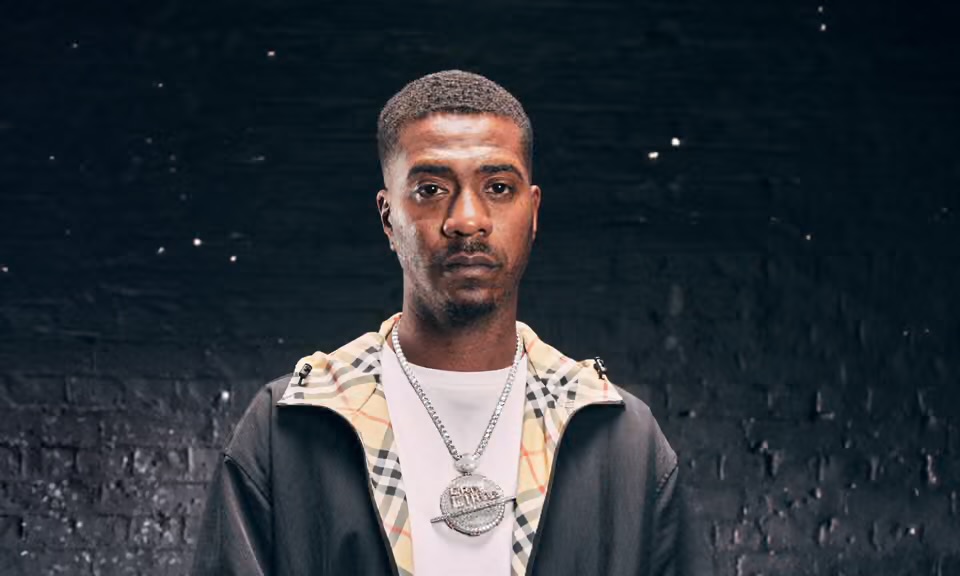 Nines photographed in a black jacket with Burberry-style check lining, silver chain pendant, against a dark charcoal brick wall.