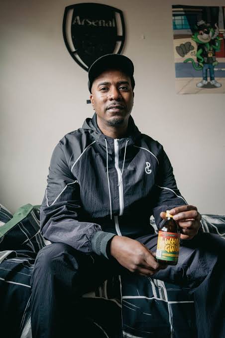 Nines sitting on a bed wearing a dark windbreaker with a DG logo, holding a small amber bottle.