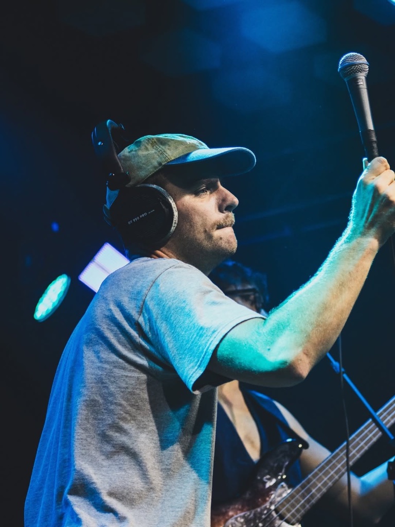 Mac DeMarco holding mic high under teal and blue stage lights