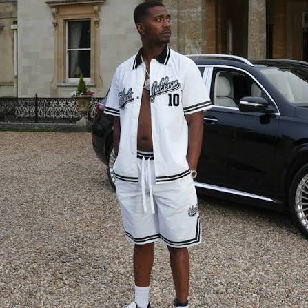 Nines in a white baseball-style shirt with 'Dolce' script, standing in a gravel driveway outside a beige stone building.