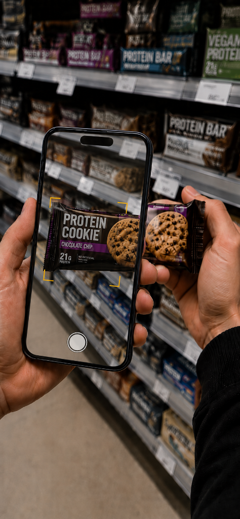 Telefon v ruce skenuje protein cookie před regálem s realistickým camera preview a tlačítkem fotoaparátu.