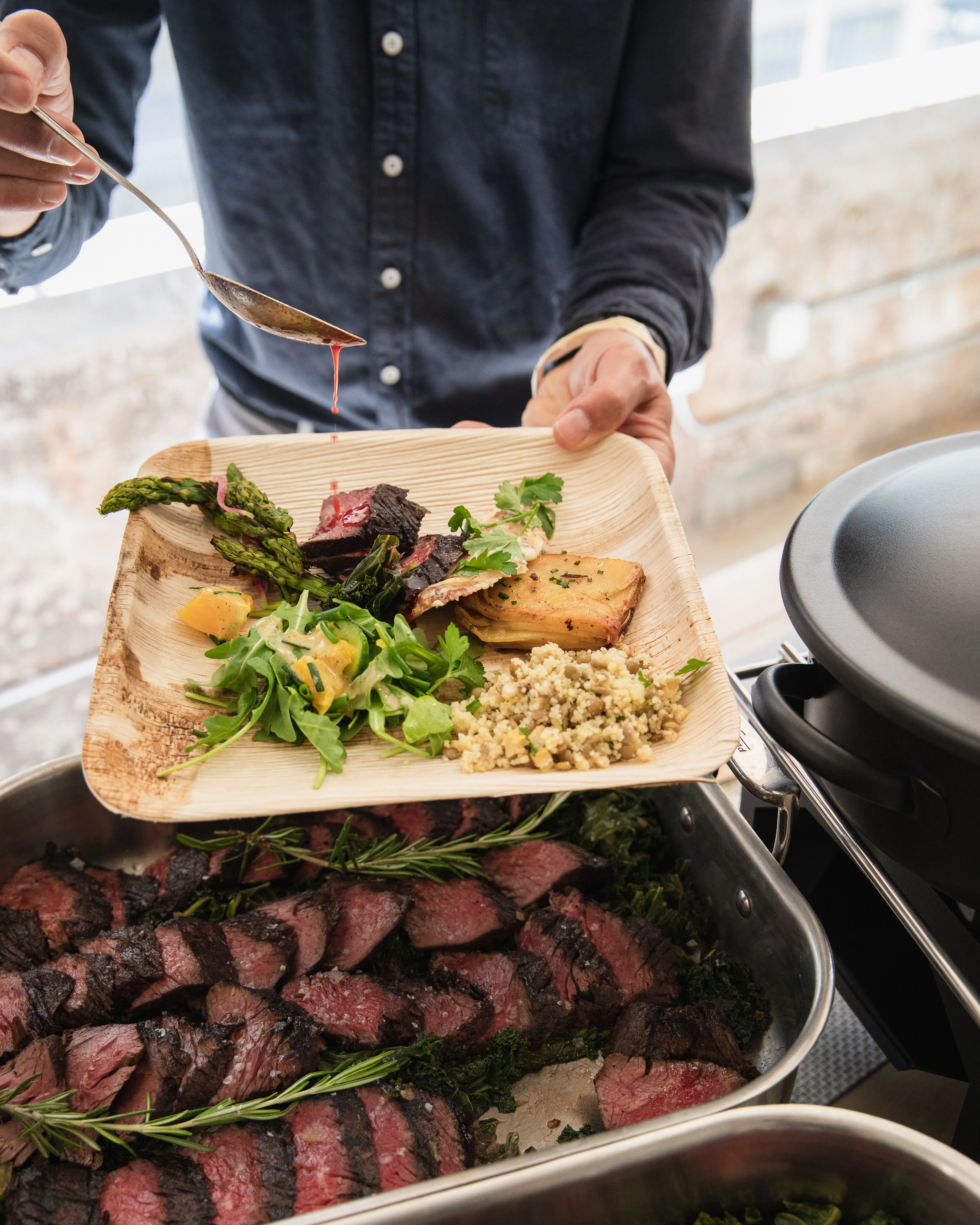 Chef drizzling red jus over seared beef, asparagus, and grain salad on a palm-leaf plate