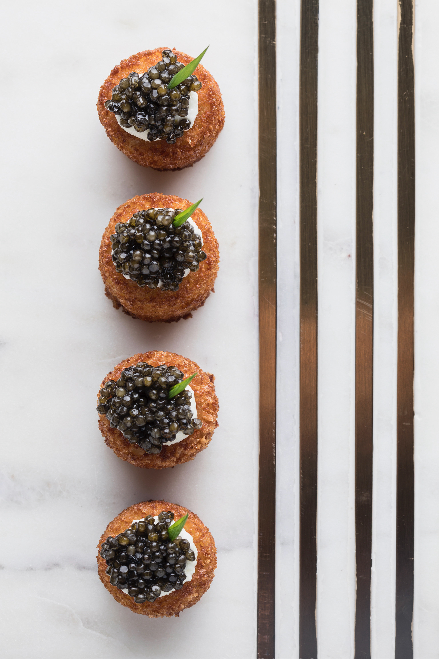 Four caviar-topped fried hors d'oeuvres on marble beside brass bars