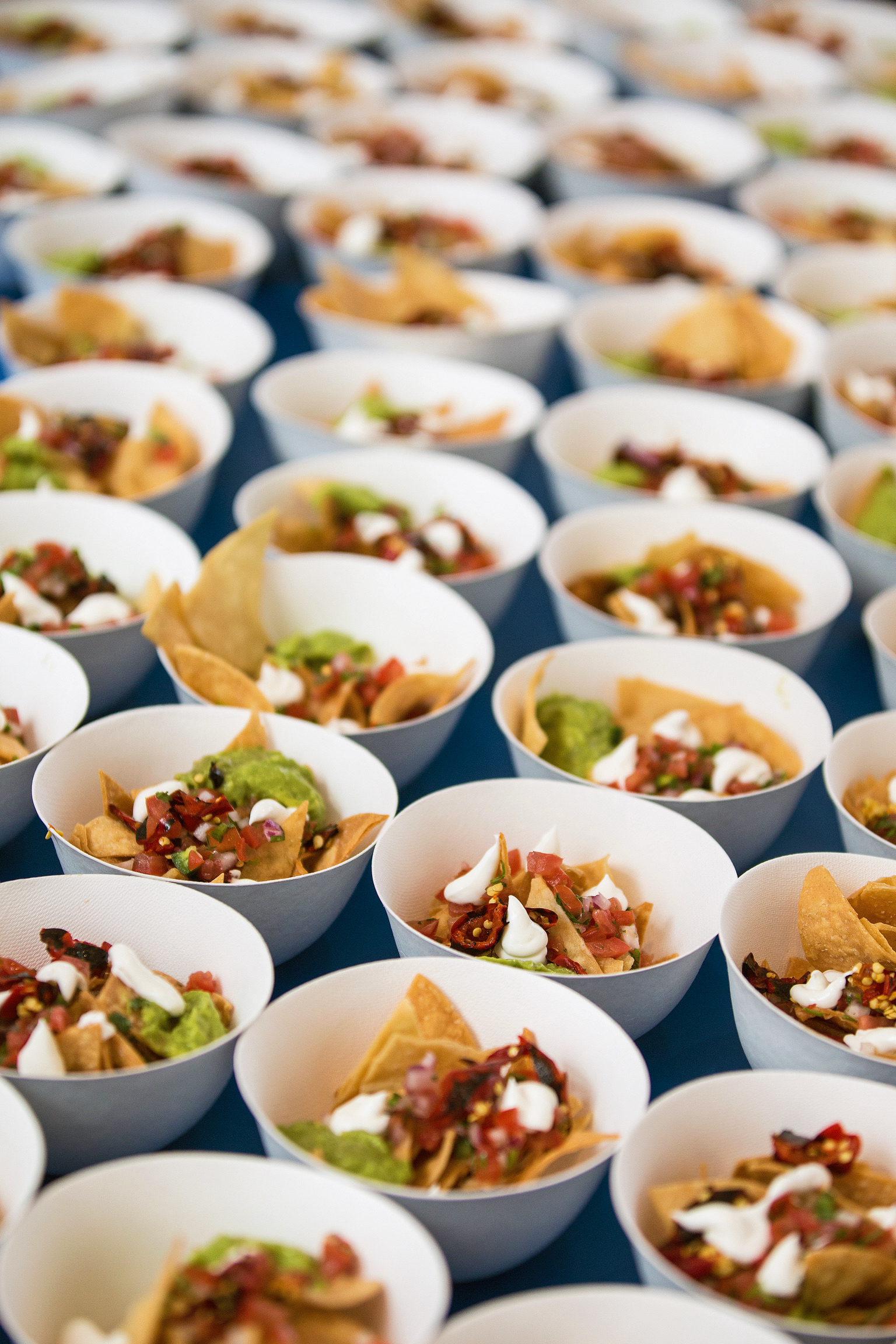 Grid of white paper bowls with tortilla chips, guacamole, salsa, and sour cream on a deep blue surface