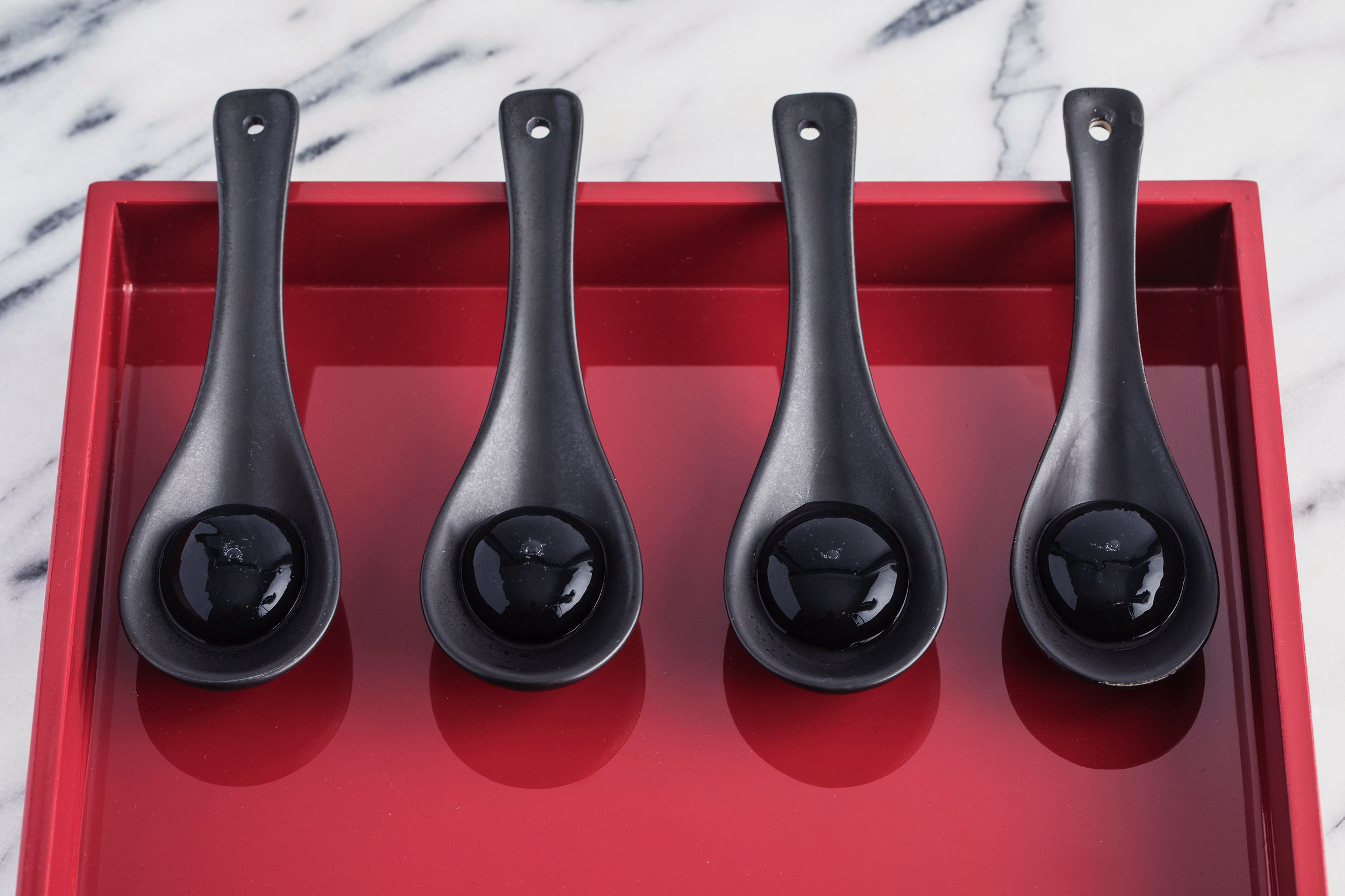 Four black molecular gastronomy spheres in ceramic spoons on a glossy red tray