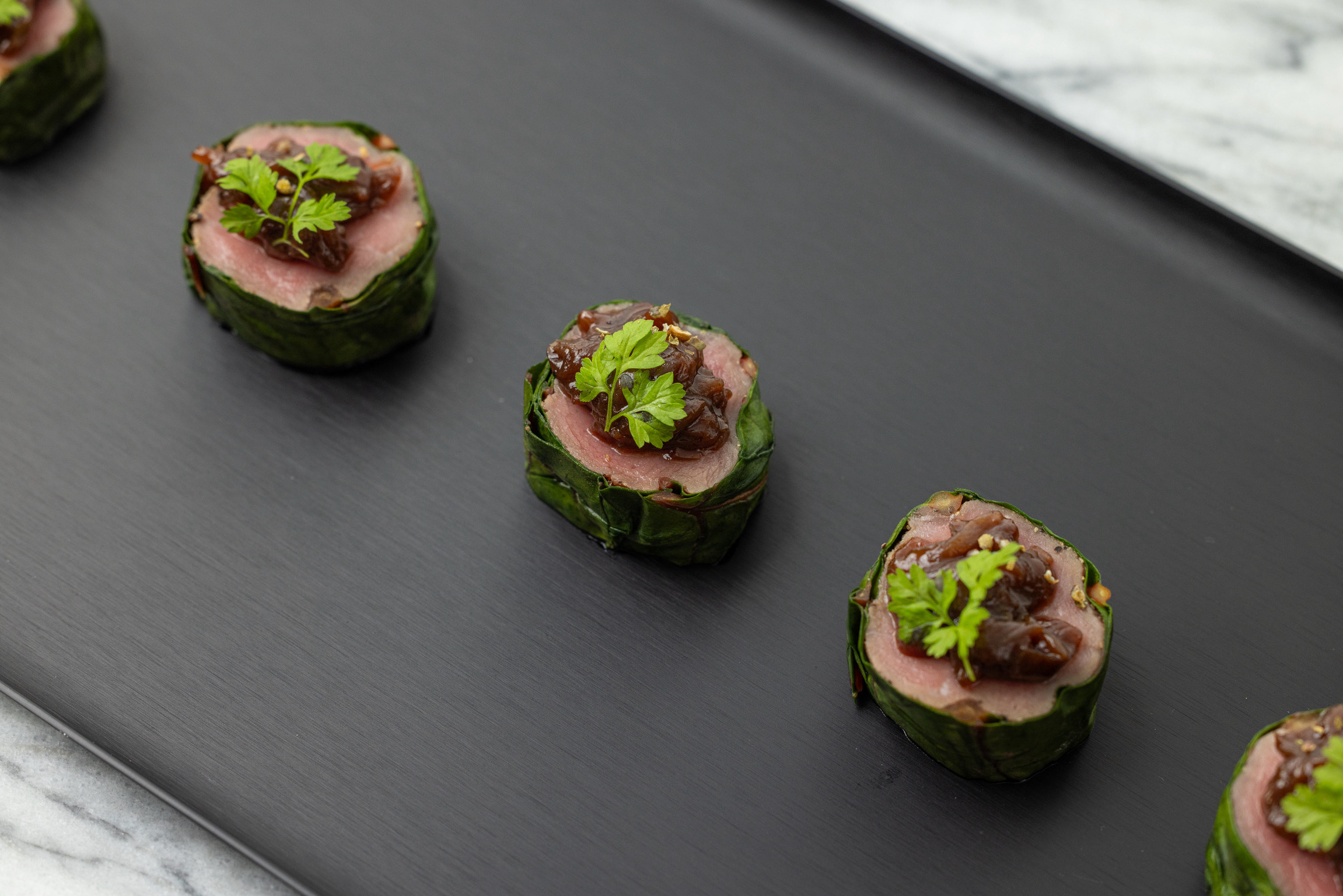Three beef and leaf-wrap bite-sized appetizers with caramelized onion on a matte black tray