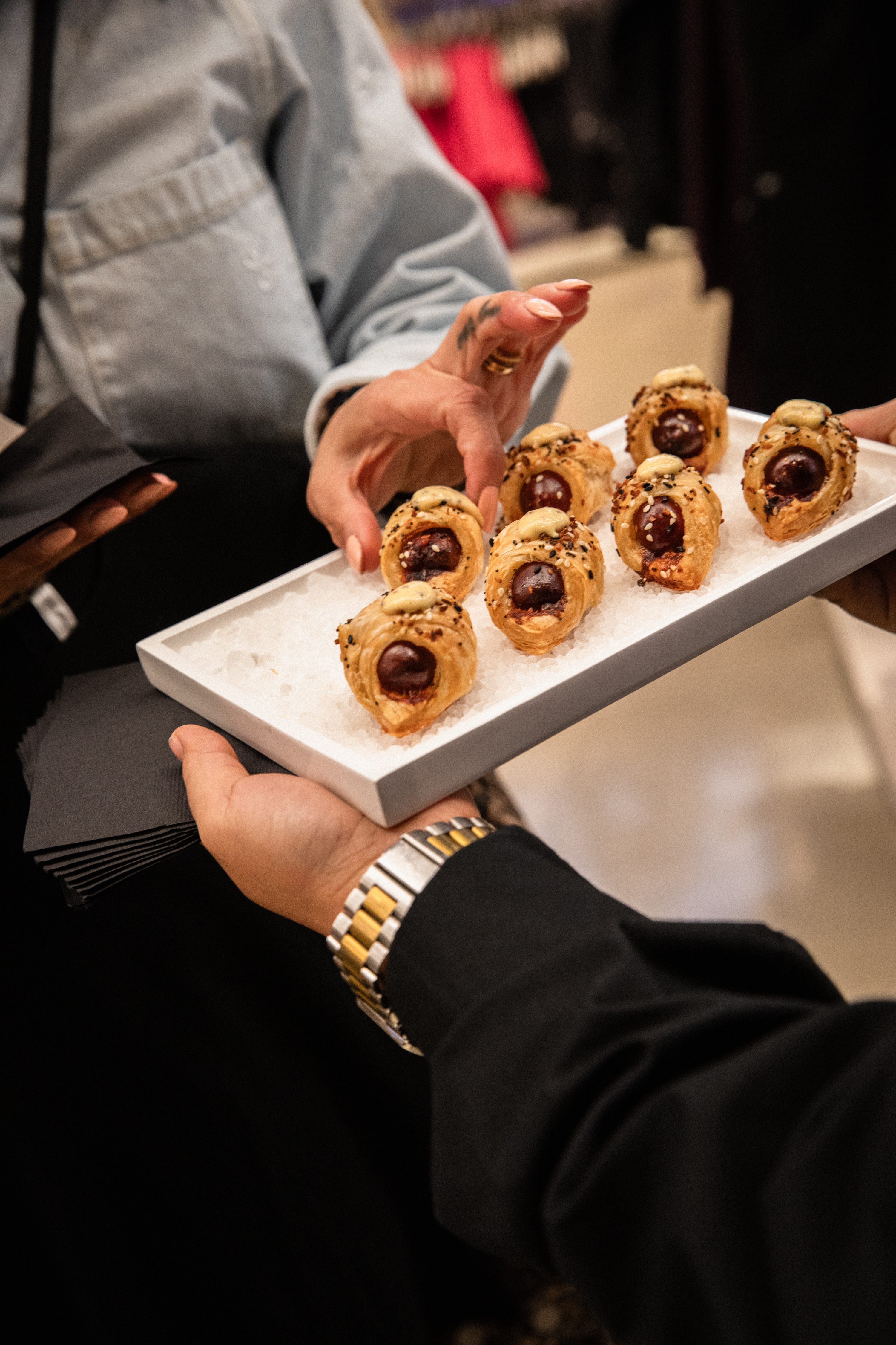 Person holding a white tray of seven flaky sausage roll pastries topped with pale sauce and sesame seeds on a bed of coarse salt
