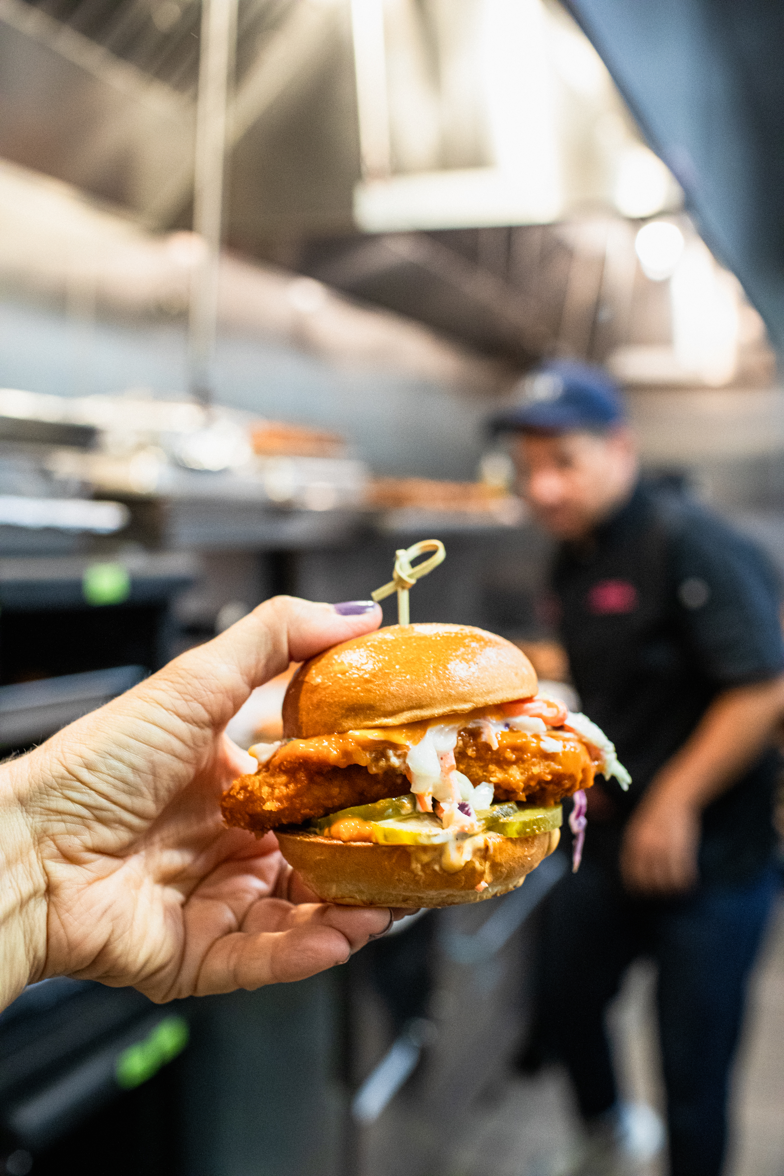 Hand holding a fried chicken slider with slaw and pickles on a toasted bun