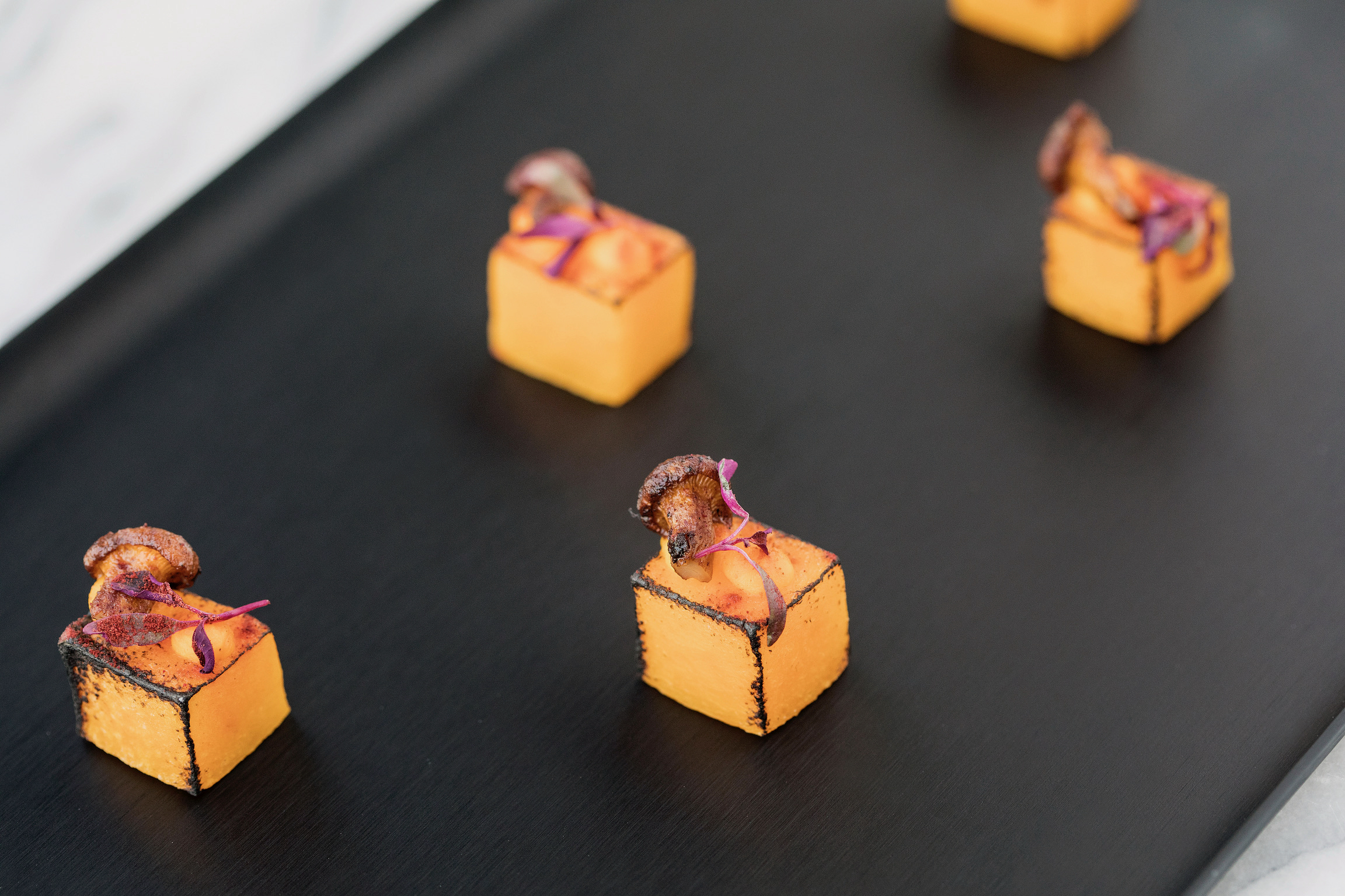Four roasted squash cubes with mushrooms and purple micro-greens on a black tray
