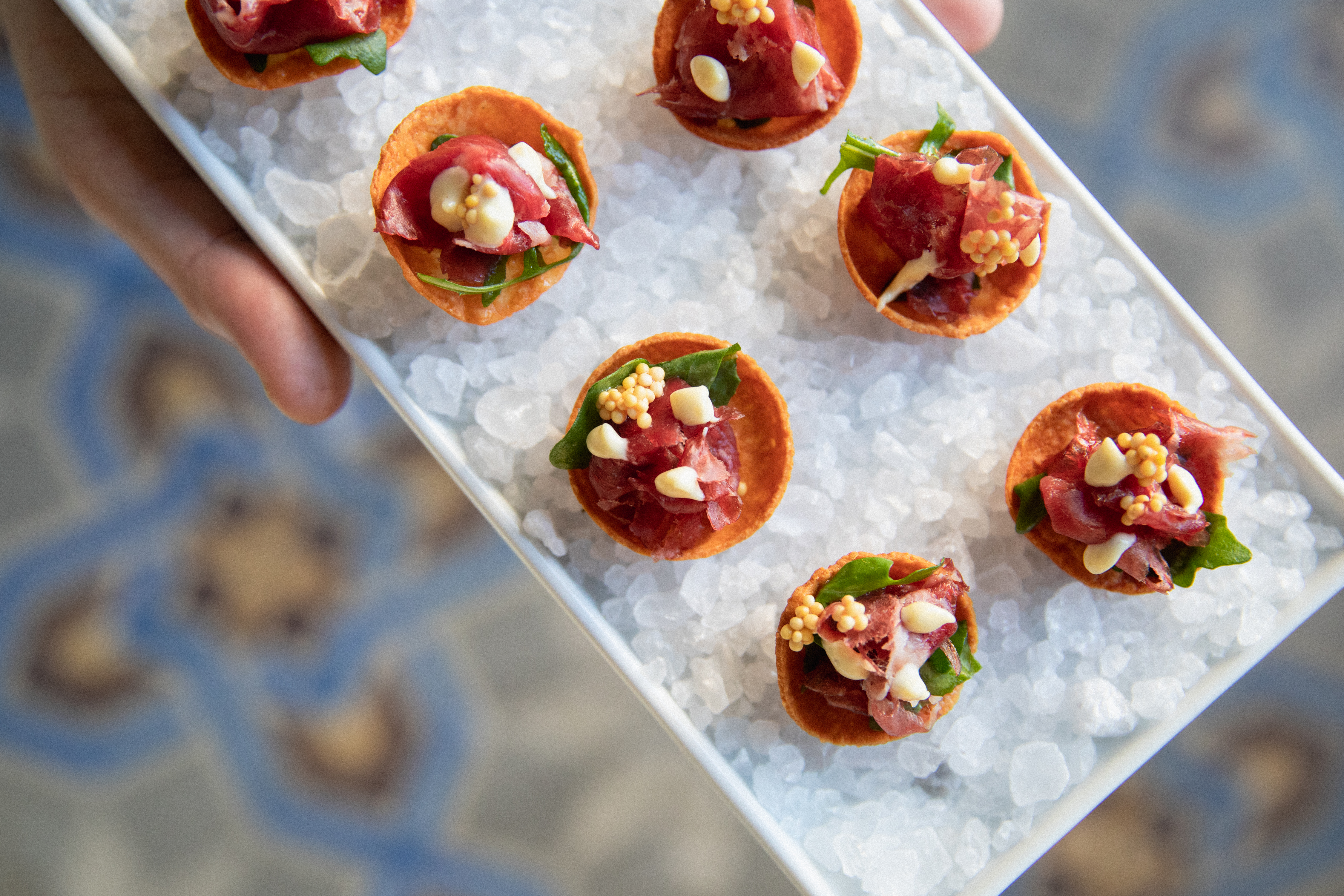 Top-down view of a white tray on coarse sea salt holding seven crispy shells filled with beef tartare, arugula, and mustard seeds