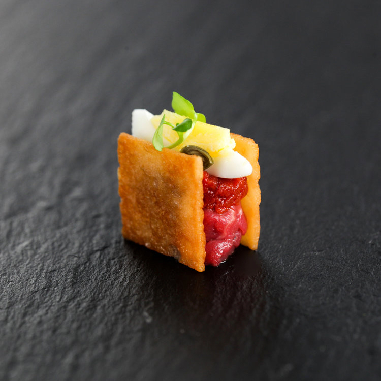 Single steak tartare bite on a crispy shell with egg white and microgreens on dark slate