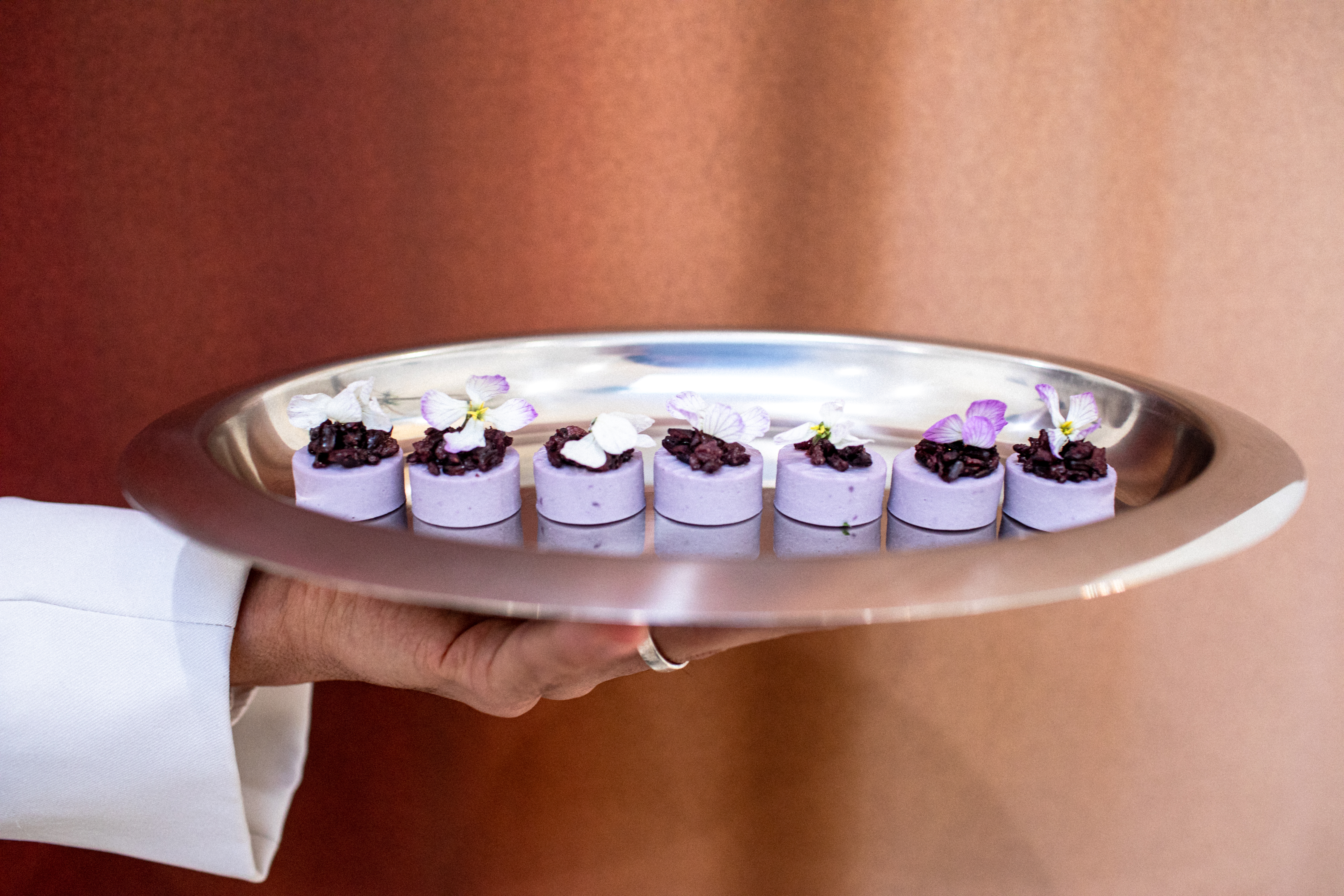 Hand holding a silver tray of seven light-purple cylindrical desserts topped with dark jam and tiny edible flowers