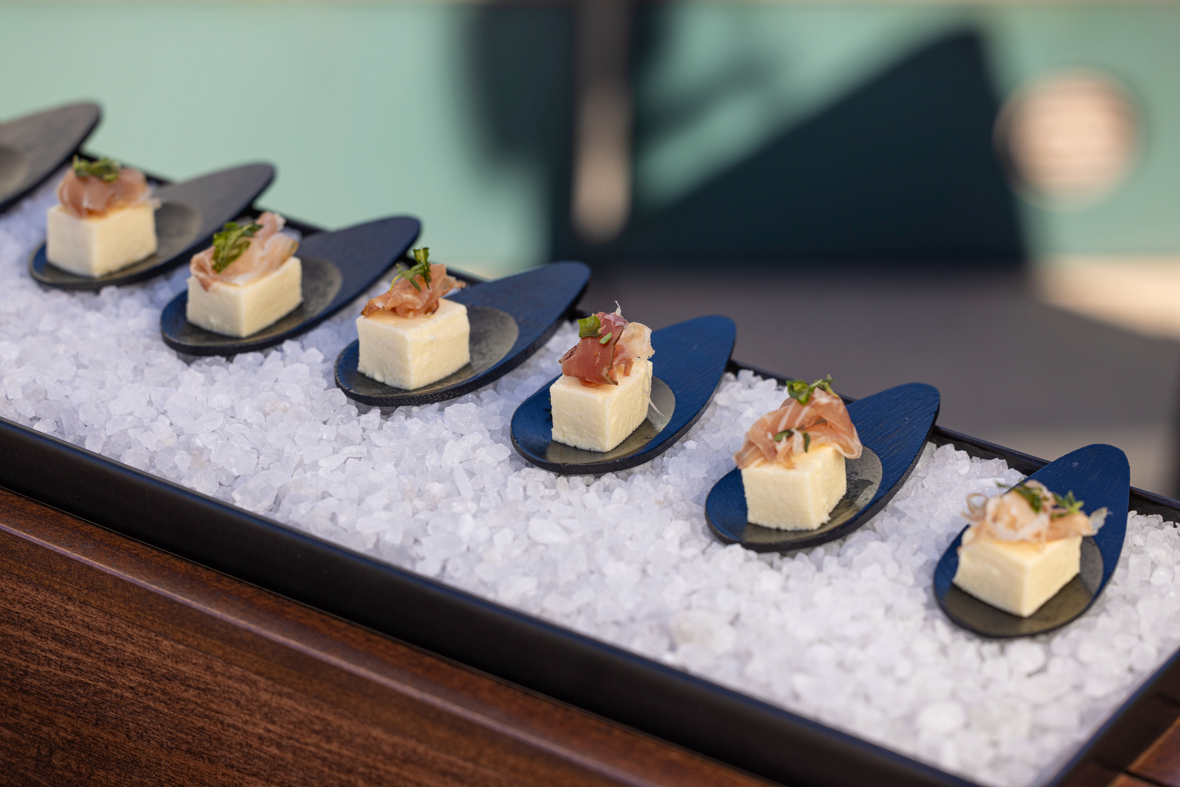 Six sweet corn panna cotta with prosciutto di Parma bites on matte black petal spoons nestled in sea salt within a wooden tray