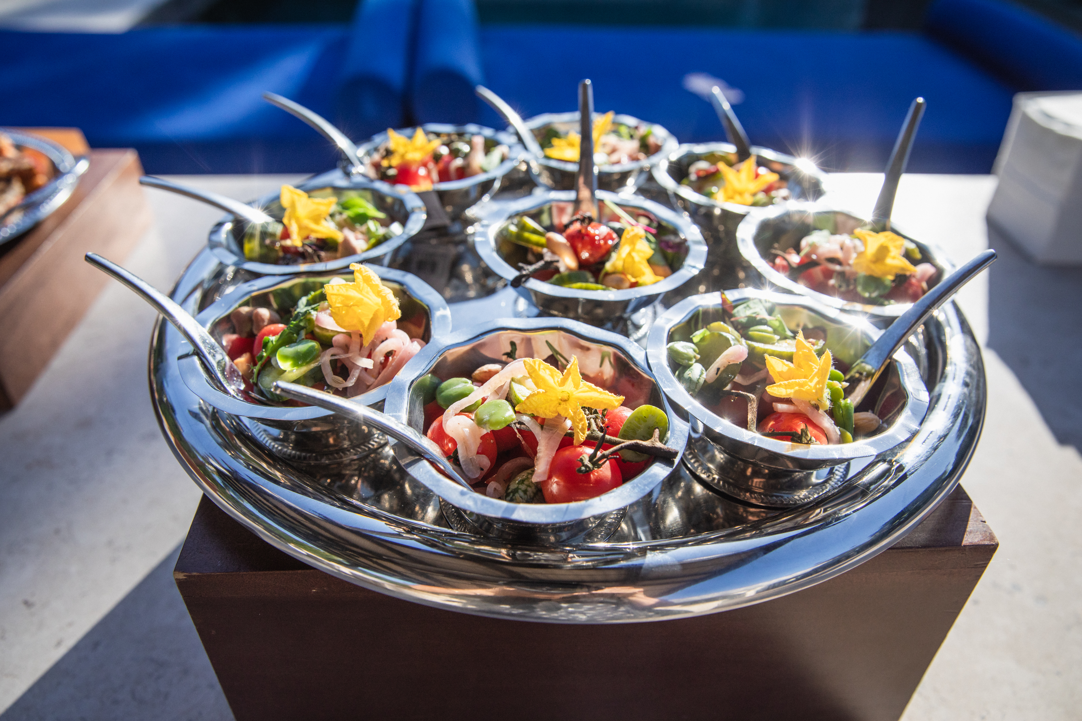 Stainless steel tray of silver bowls filled with cherry tomato and fava bean salad topped with yellow edible flowers