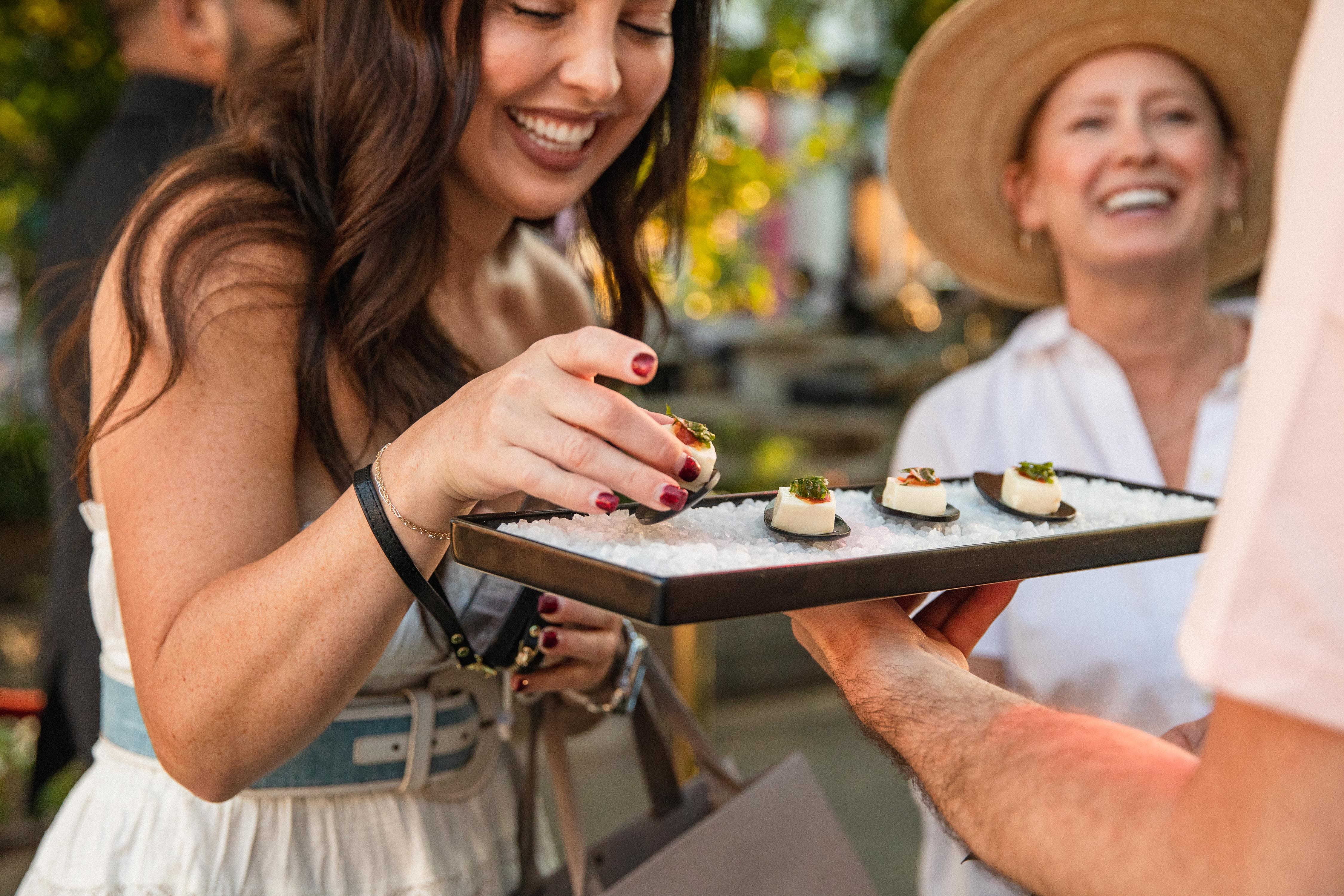 Guest reaching for a white hors d'oeuvre from a salt-bed tray at an outdoor cocktail event