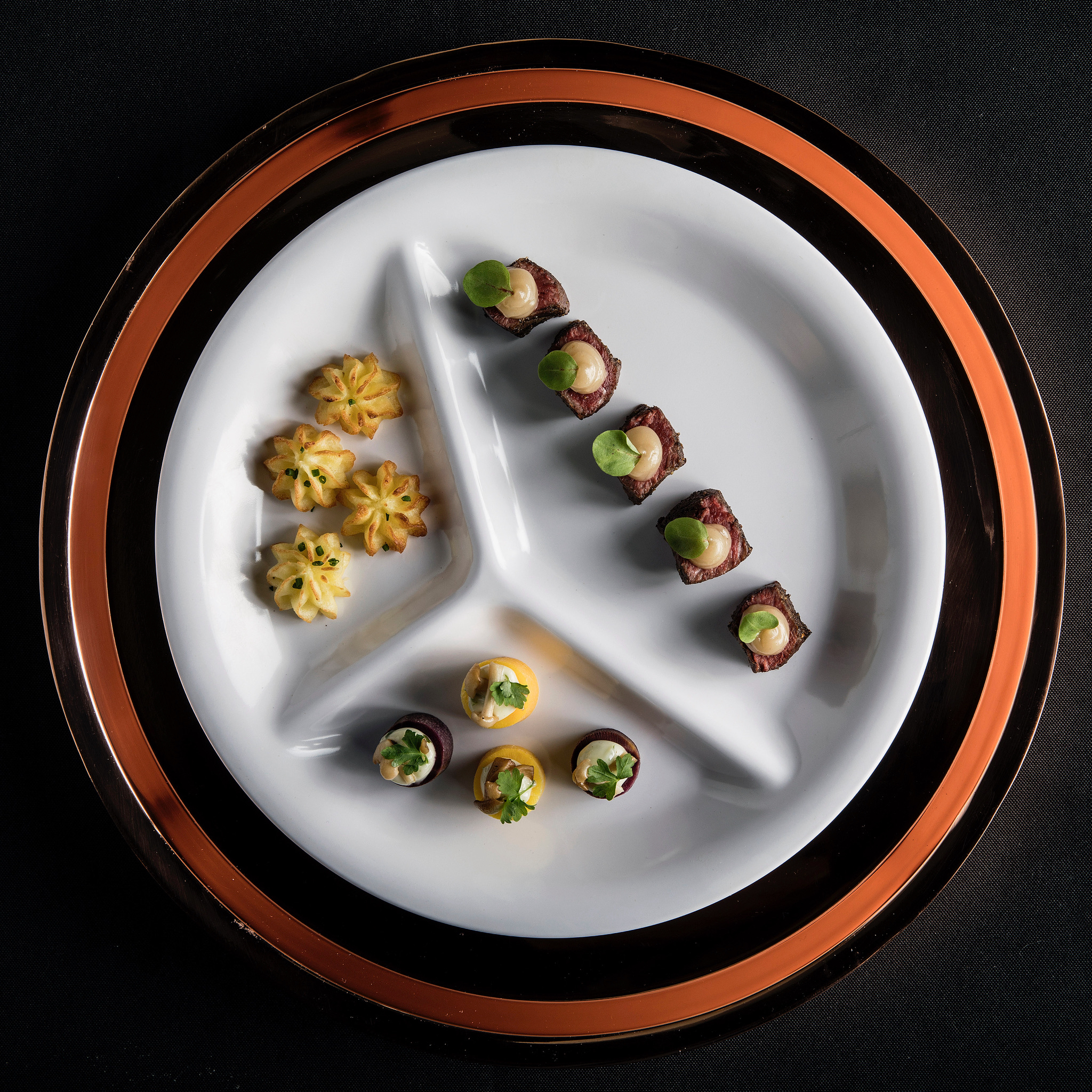 Three-sectioned catering plate with steak cubes, piped potato rosettes, and root vegetables on a copper-rimmed charger