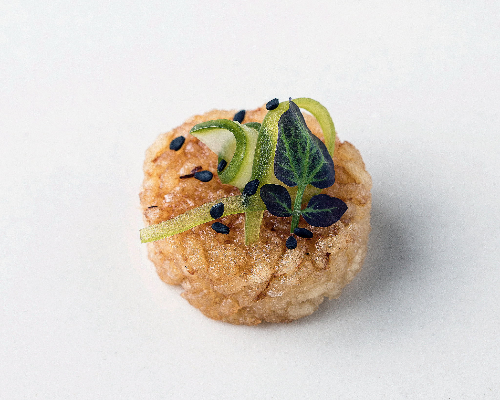 Single crispy seared rice cake topped with a cucumber ribbon, microgreens, and black sesame