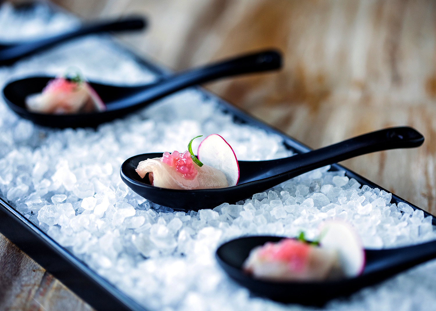 Three black tasting spoons with hamachi crudo, pink roe, and radish on a bed of sea salt