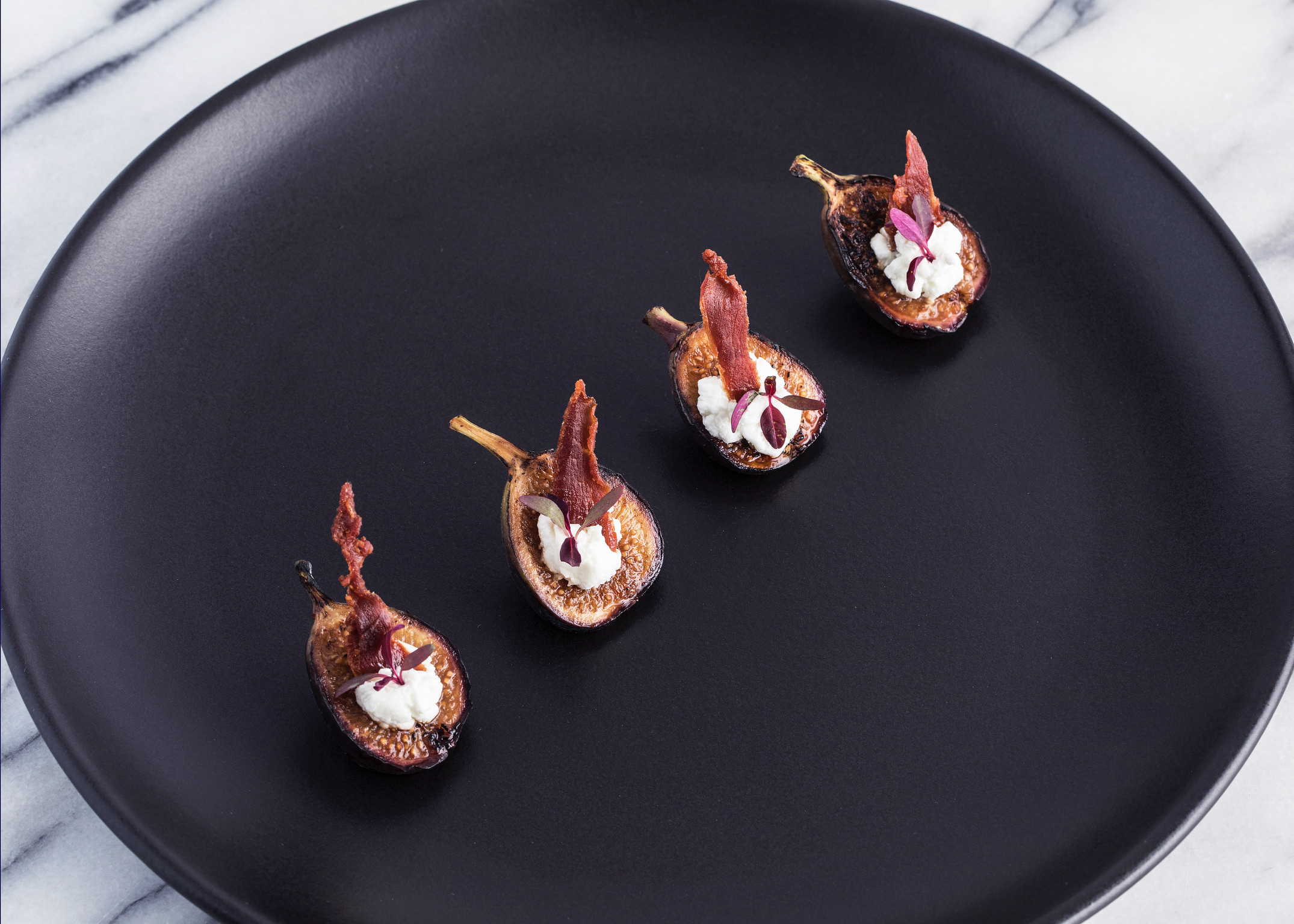 Four roasted fig halves with goat cheese, bacon, and purple microgreens on a matte black plate