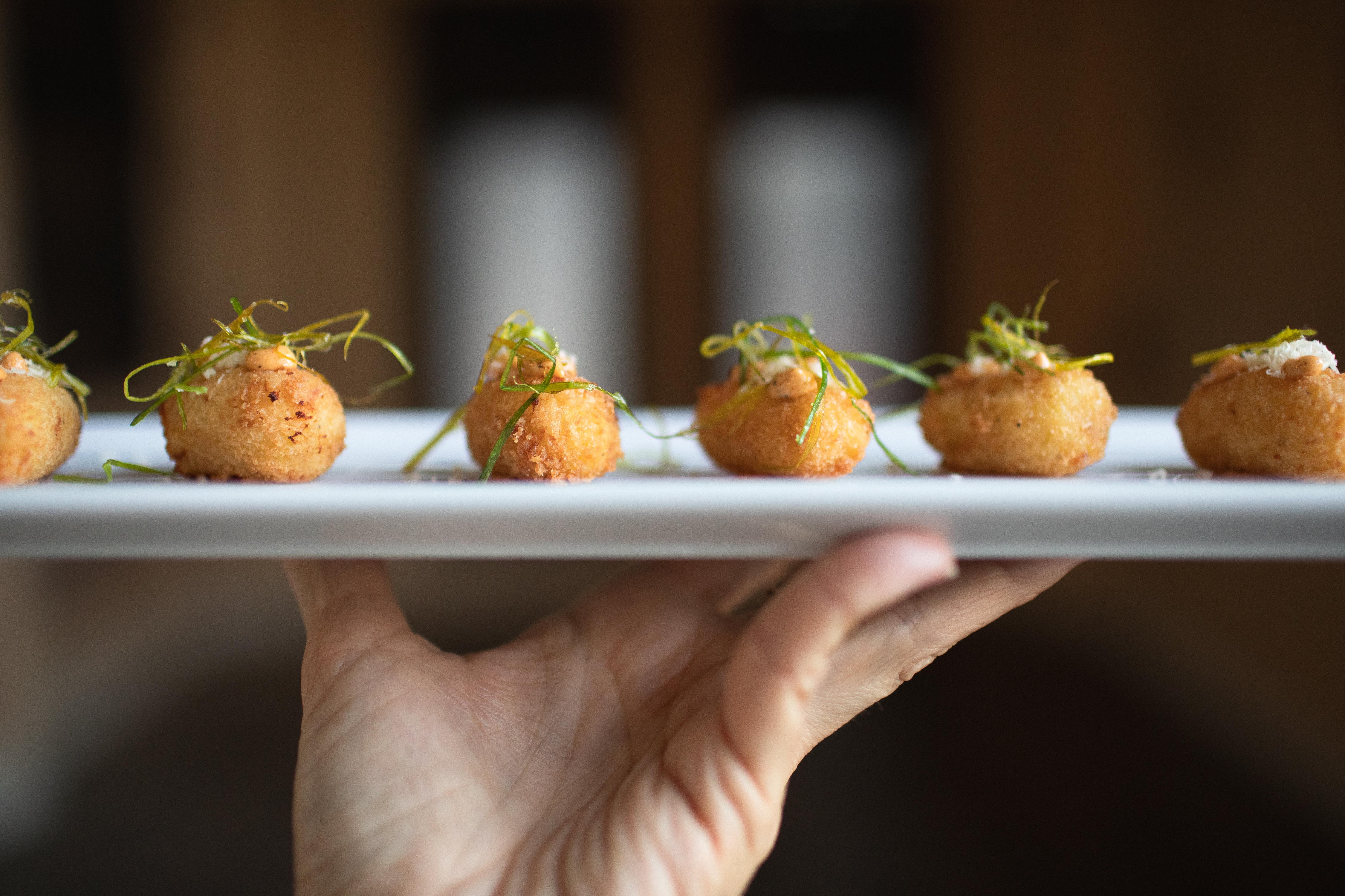 Hand holding a platter of five golden croquette appetizers topped with orange sauce and fried herbs