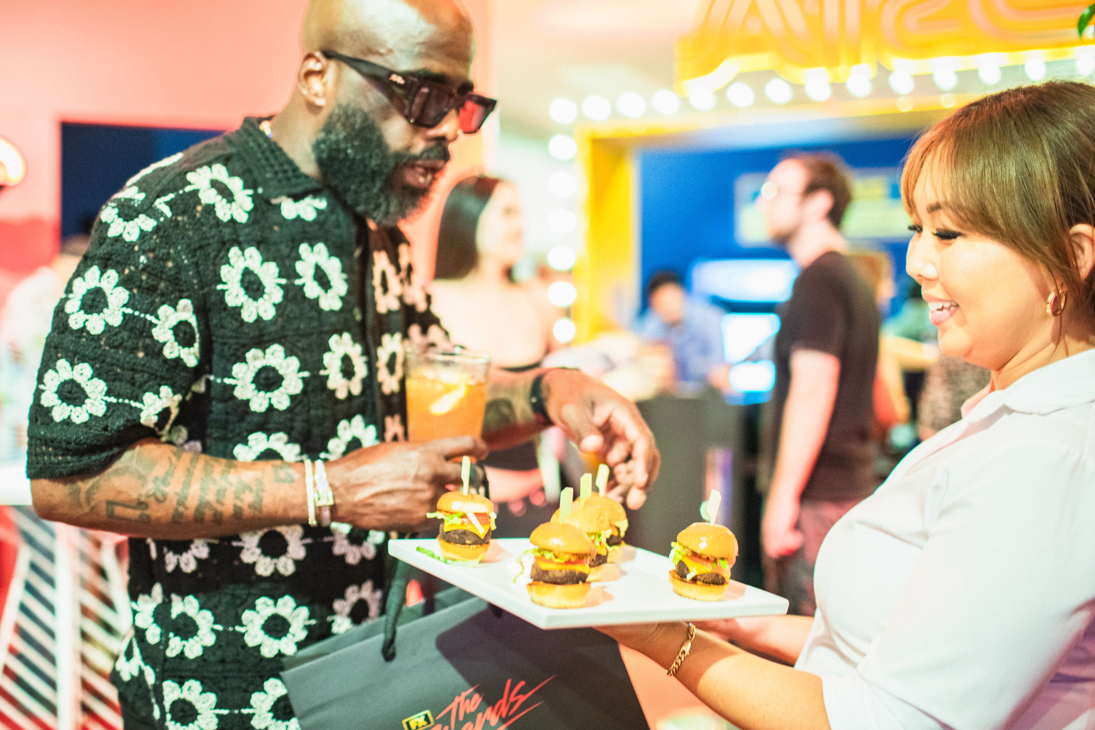Guest reaching for a slider from a white platter held by a server at a warm-toned event with a yellow-lit photo booth