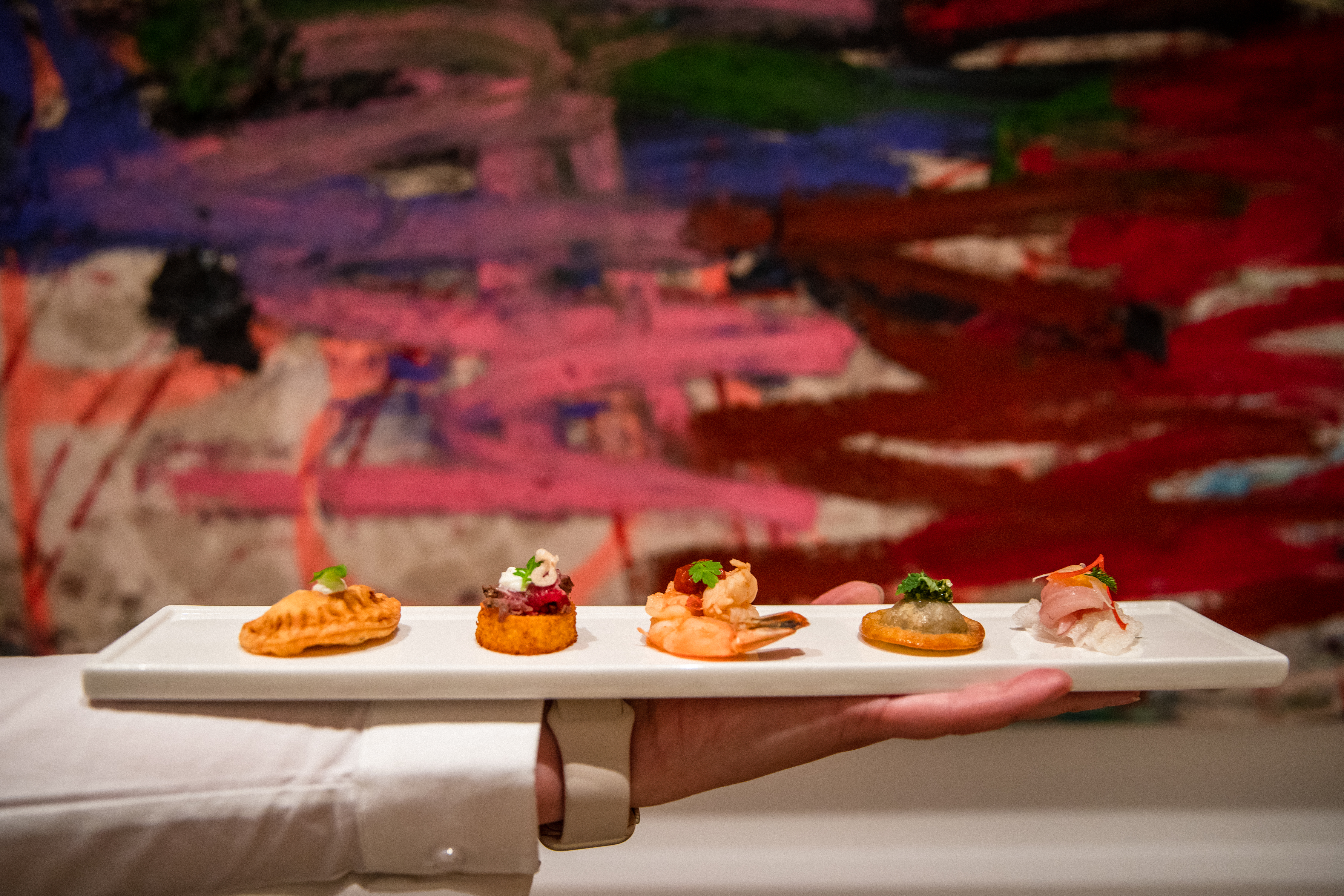 Hand holding a platter of five varied hors d'oeuvres including an empanada, crab cake, and glazed shrimp