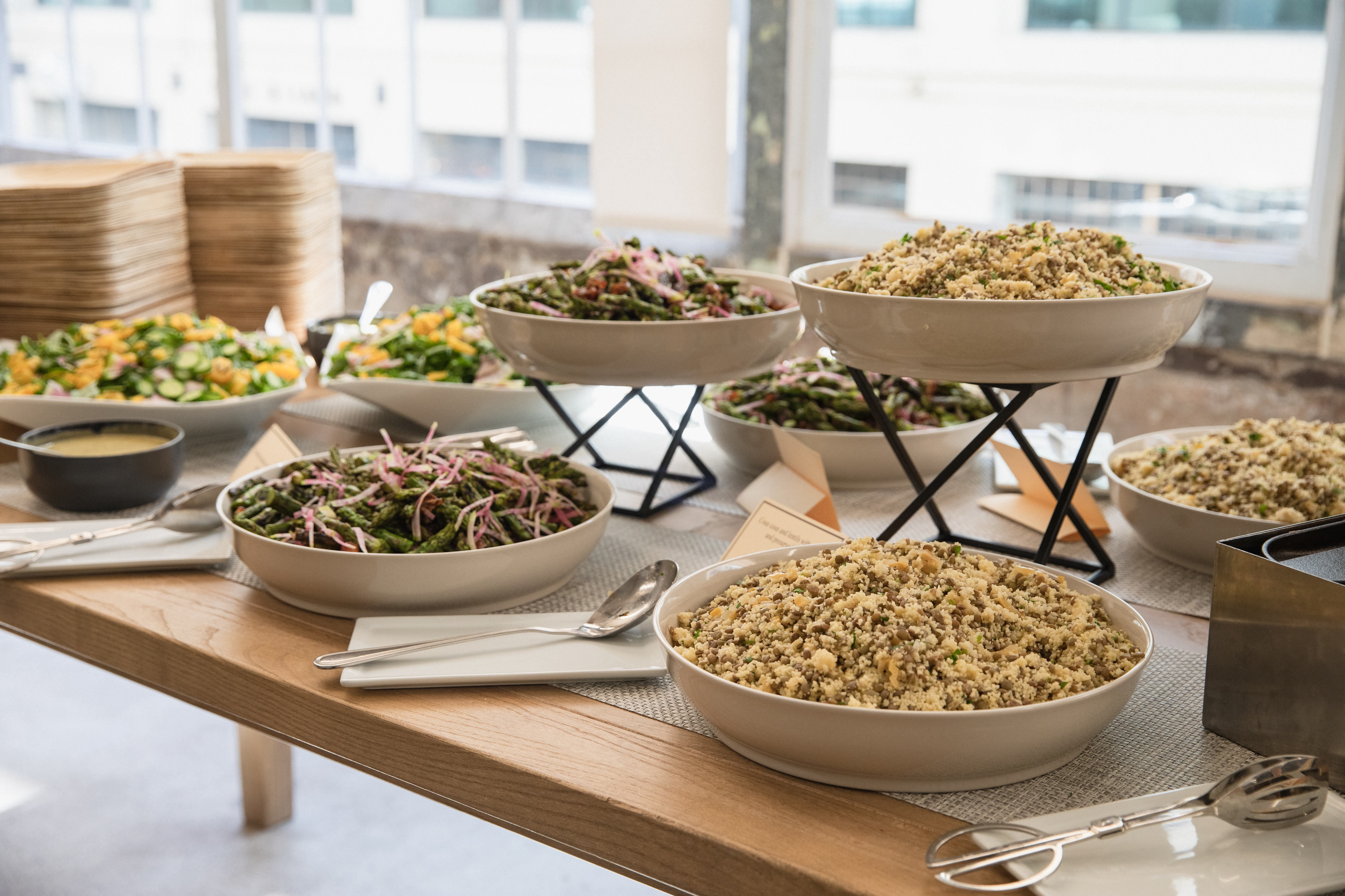 Buffet-style spread of salads and grains in white ceramic bowls on elevated stands at a catered event