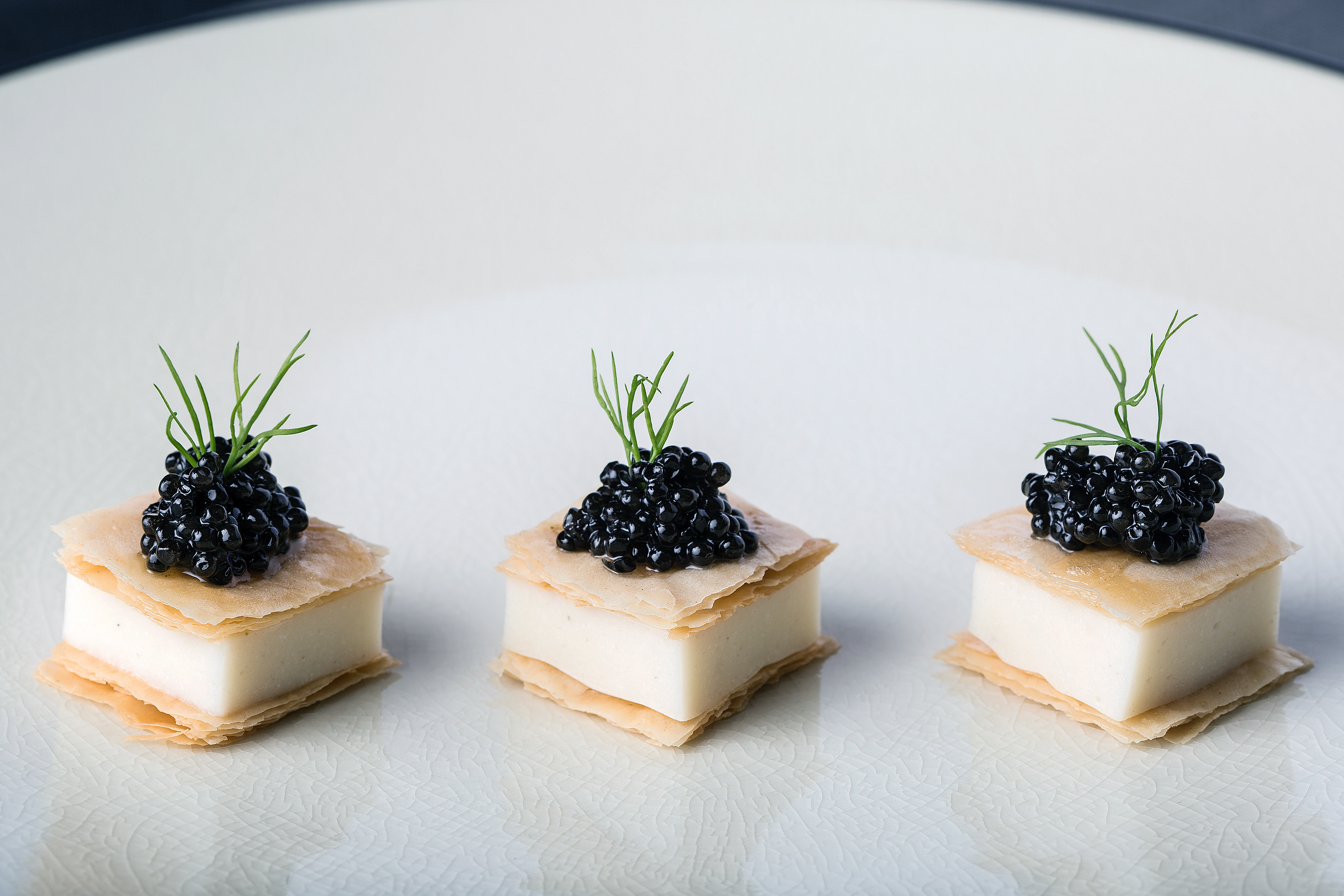 Three elegant hors d'oeuvres with white panna cotta base, golden phyllo, black caviar, and fresh dill on a cream ceramic plate
