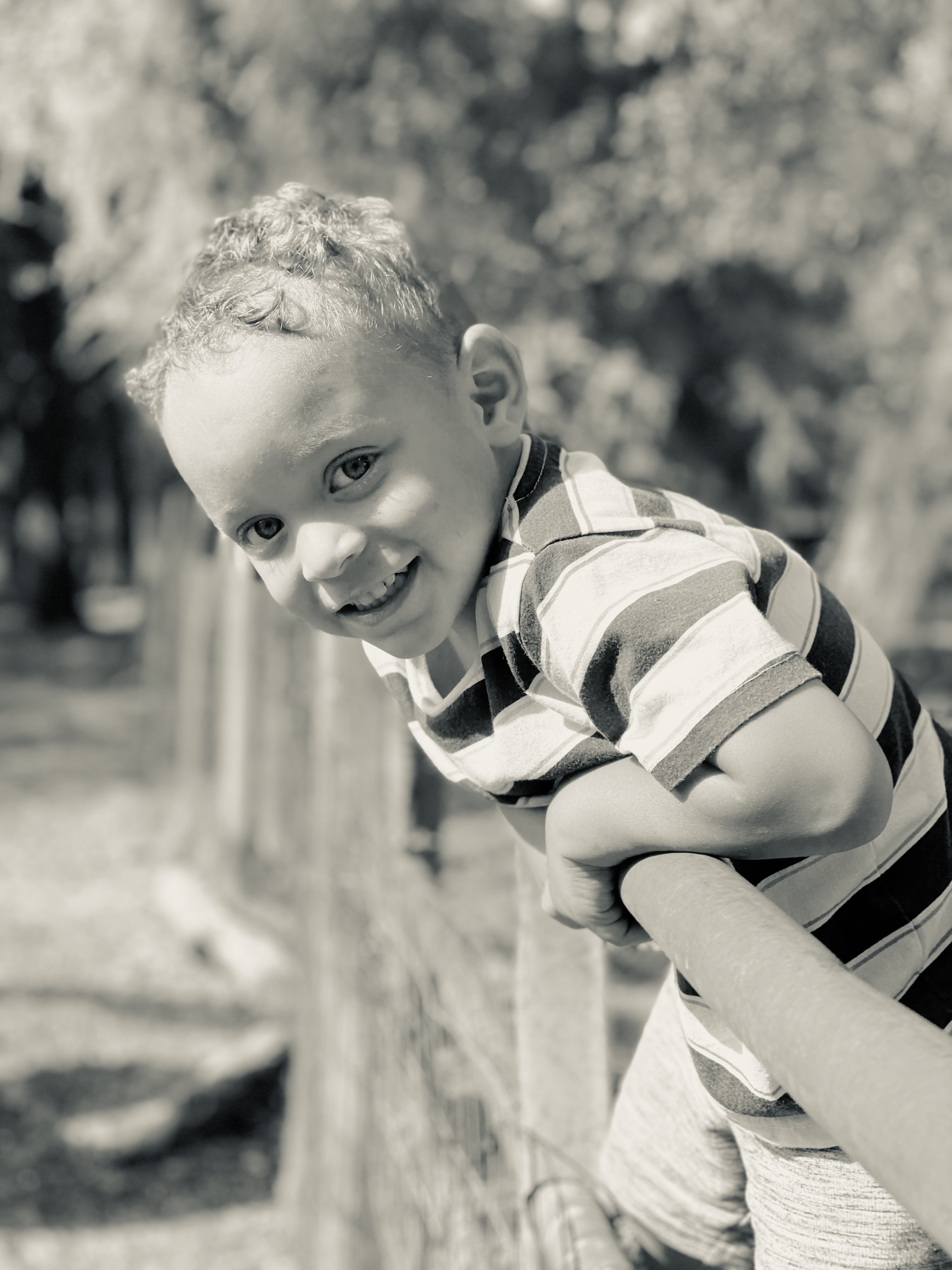 A black-and-white portrait of a curly-haired boy leaning on a metal gate, head tilted with a soft smile
