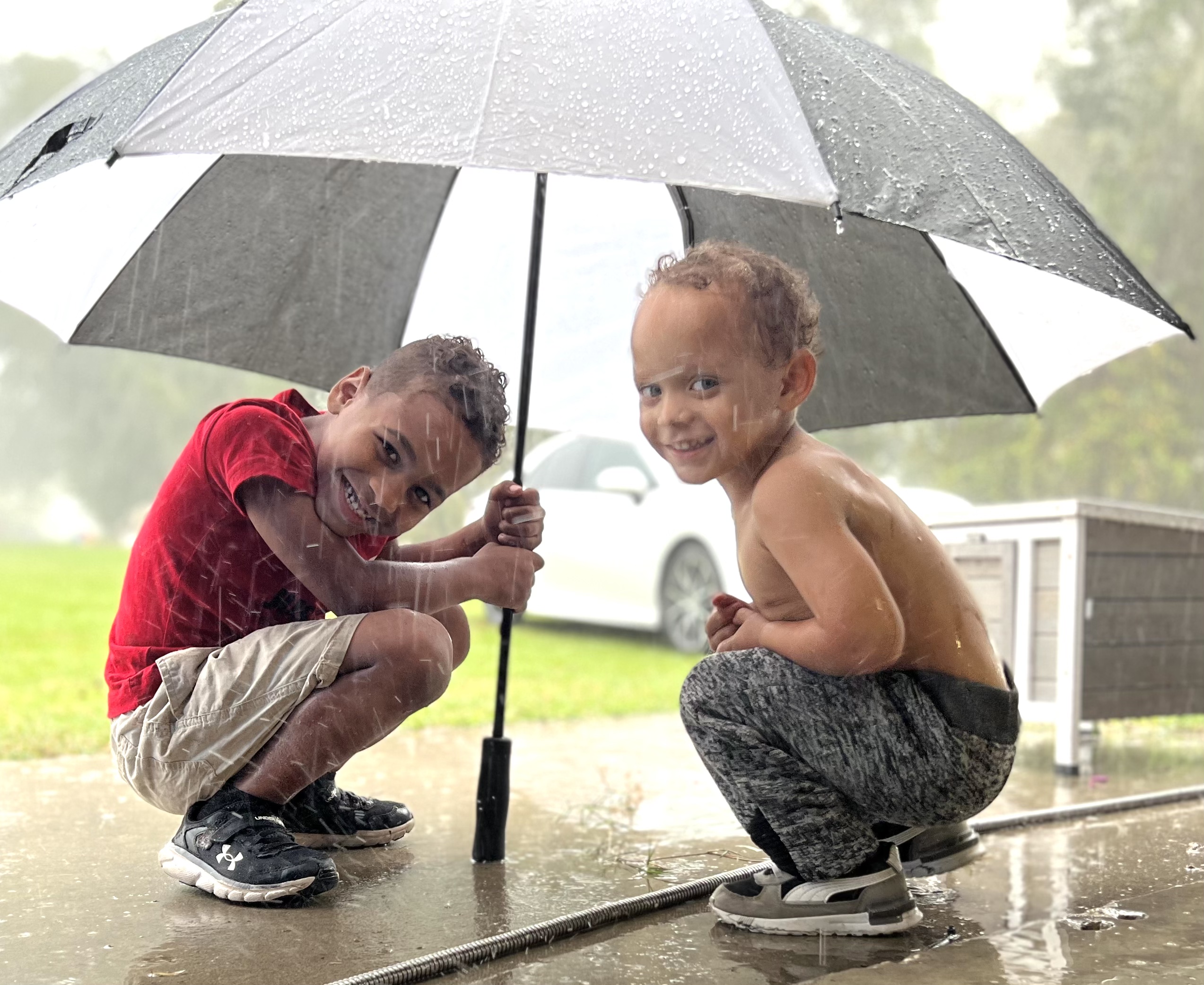 Two boys crouching under a black-and-white umbrella in the rain, both grinning at the camera on the wet concrete