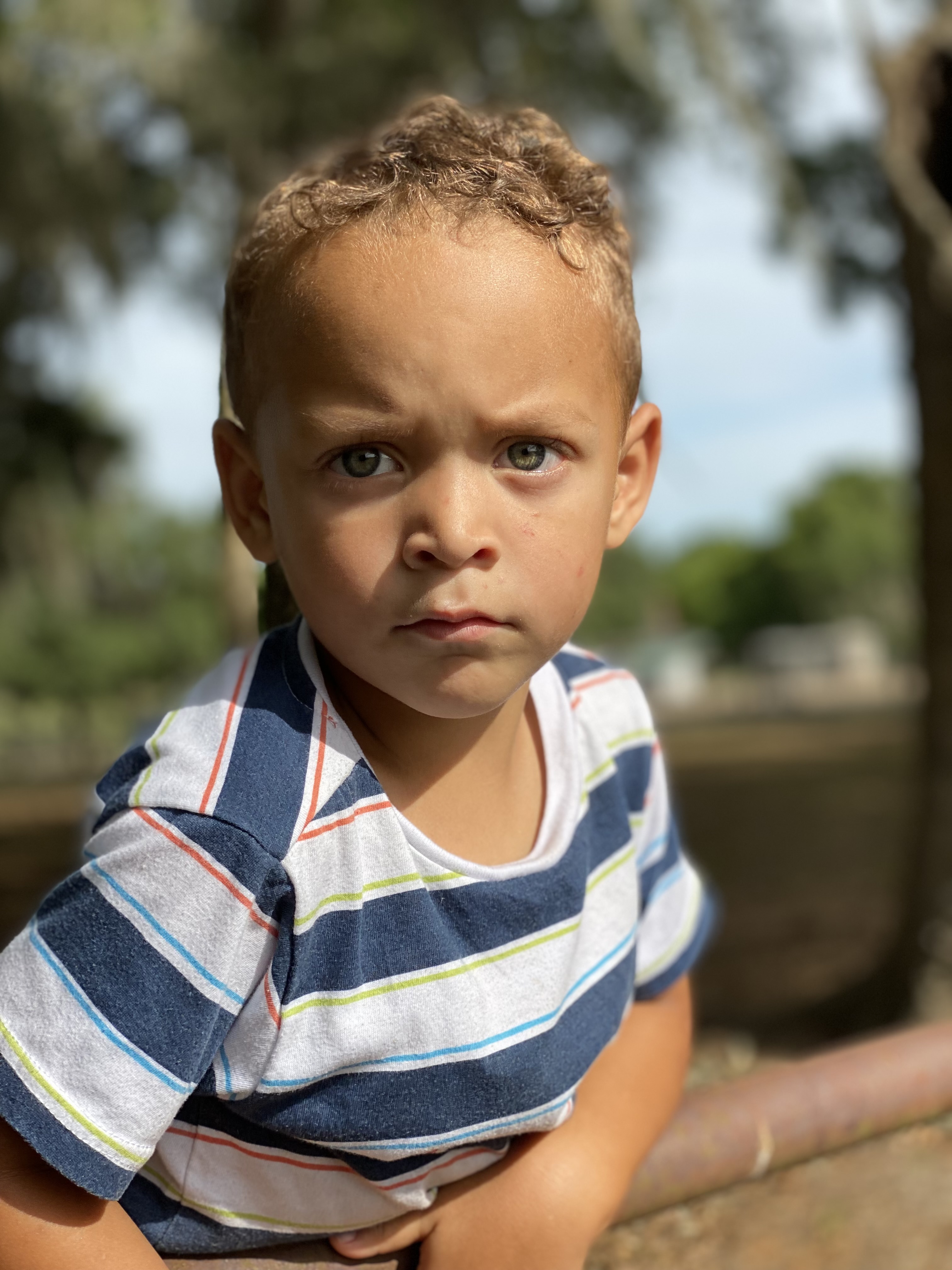 A curly-haired boy resting his hands on the rusty fence rail, facing the camera with a quiet, steady gaze