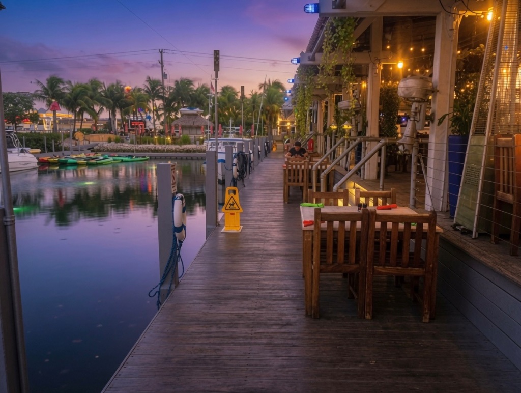 Dusk on a Key West pier — tables set for dinner under warm string lights, a yellow wet-floor sign and a life ring by the water, the sunset sky going purple and orange
