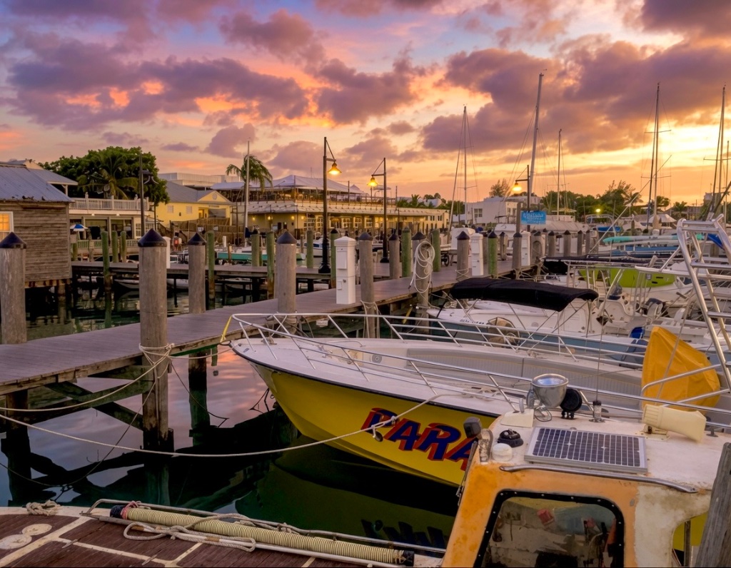 A Key West marina at dusk — sailboats and motorboats moored along the wooden pier beneath a vivid orange and violet sunset