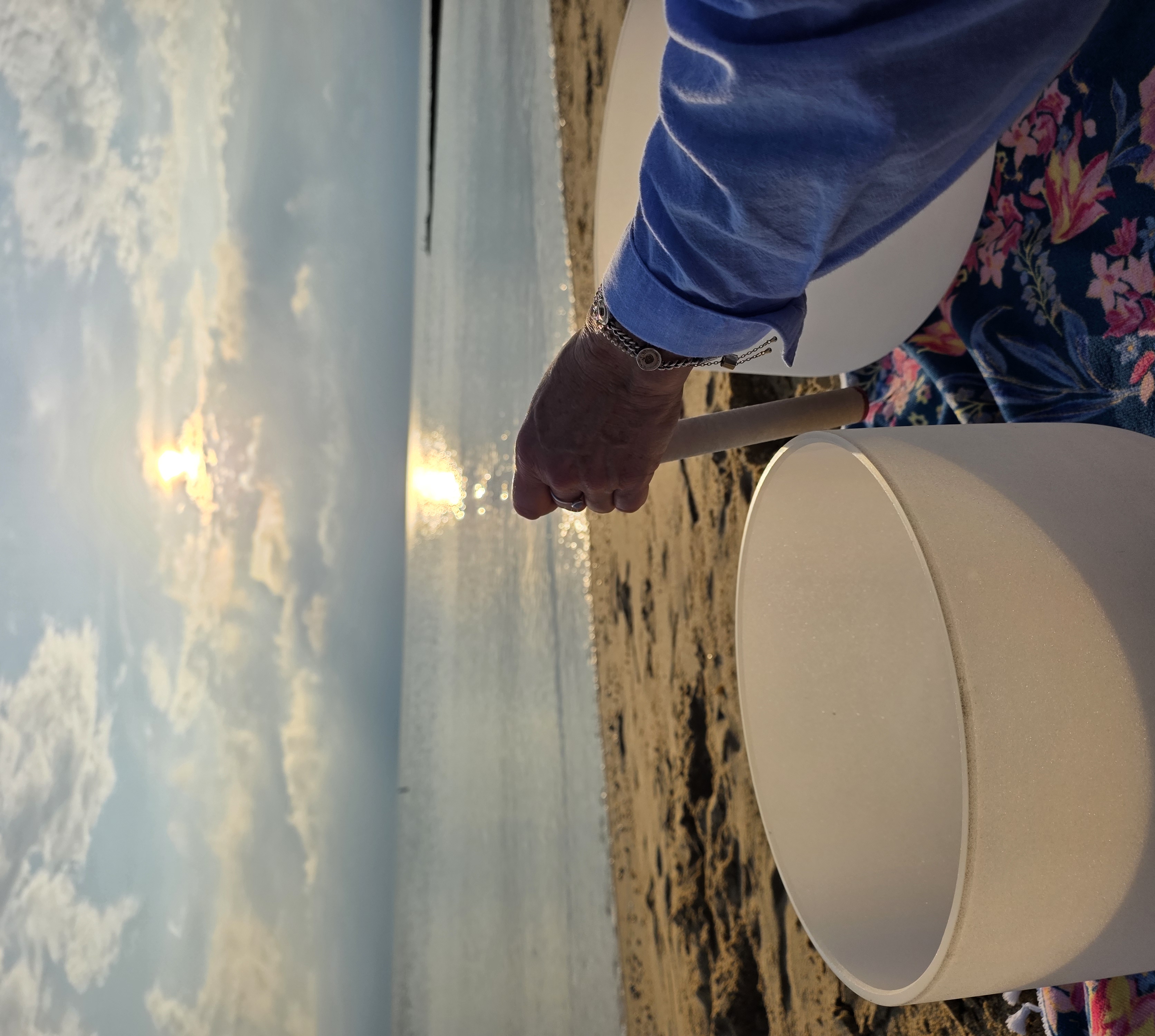 A hand playing a quartz crystal singing bowl on the sand as the sun rises over calm water
