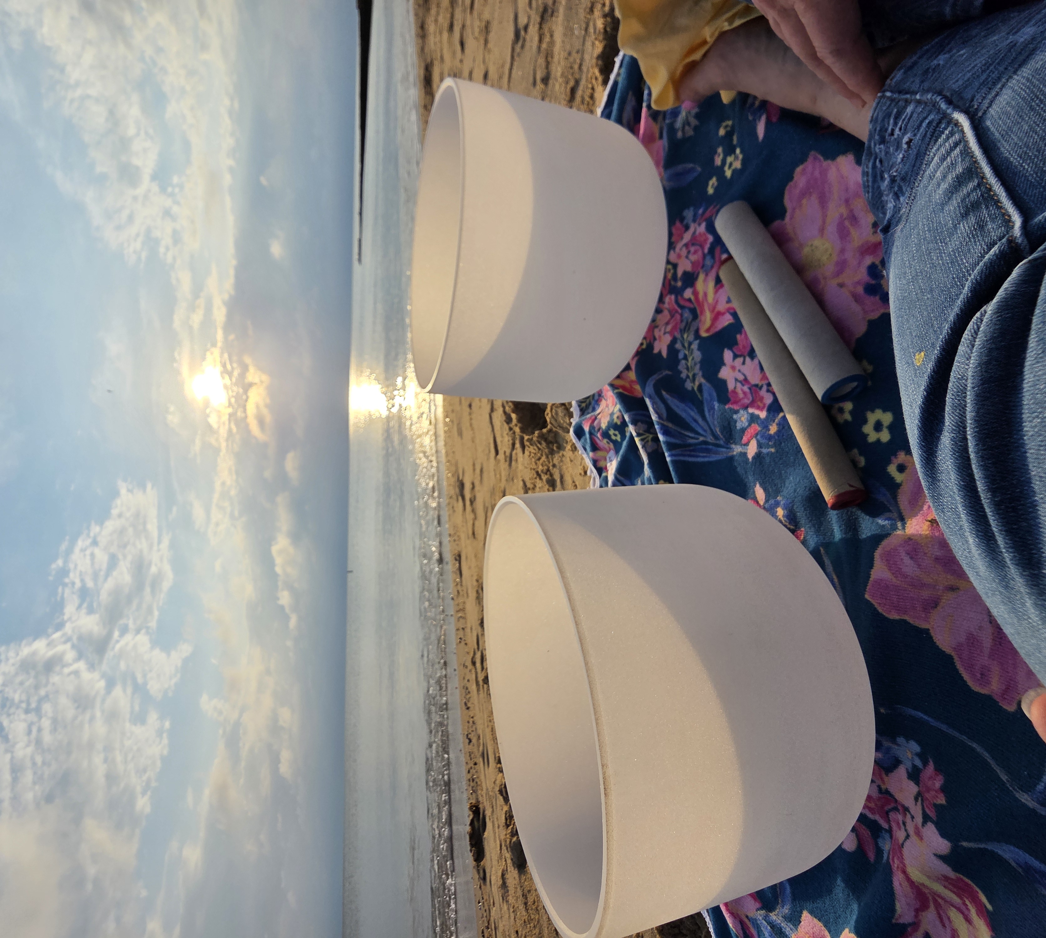 Two quartz crystal singing bowls resting on a floral cloth at the shoreline with a striker beside them at sunrise