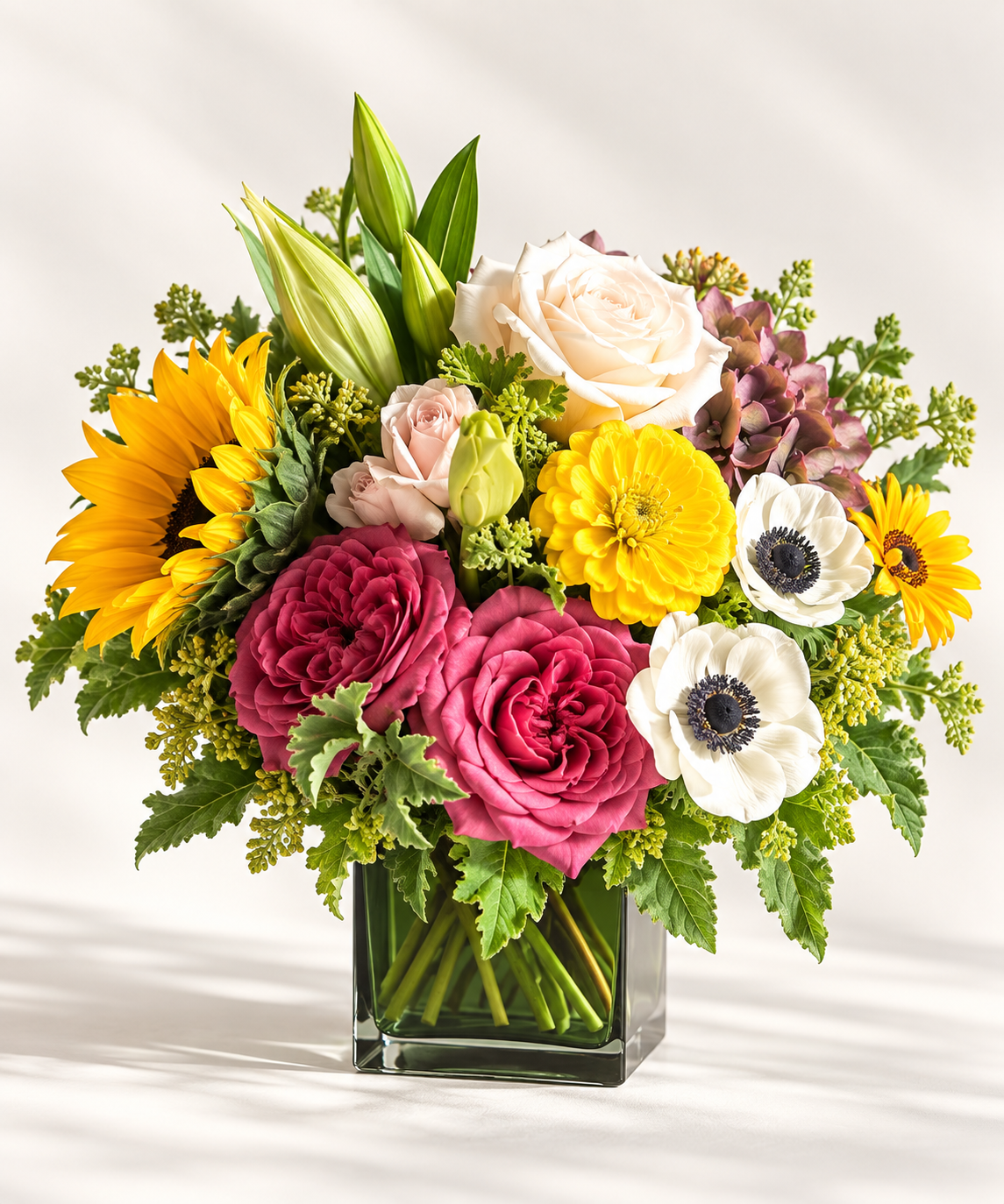 standard: Vibrant magenta garden roses, yellow sunflower, white anemones with dark centers, and purple hydrangea in a clear square glass vase — custom new baby flower delivery to Las Vegas hospitals by Bud Weismiller.