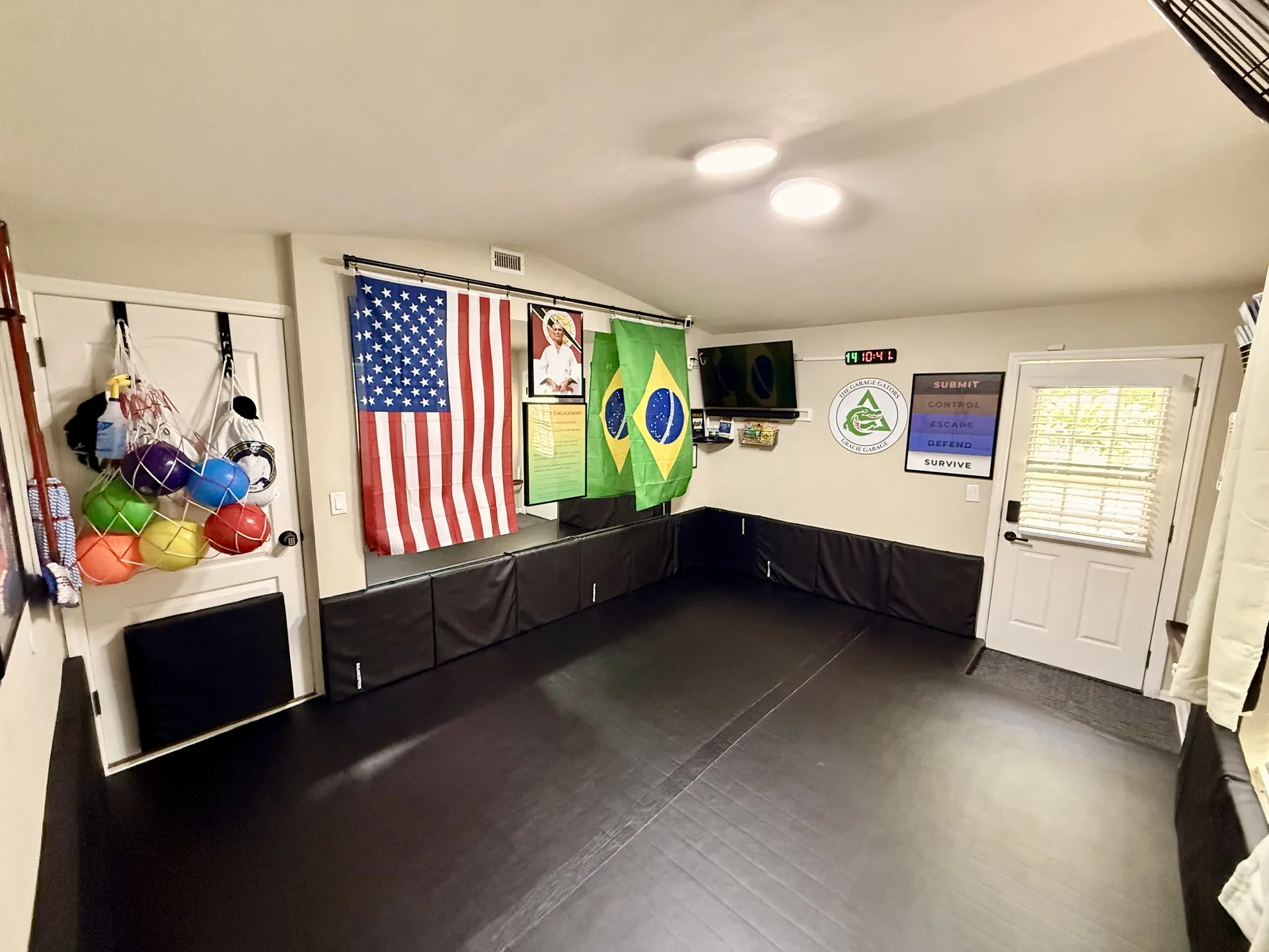 Interior of the Garage Gators Gracie Garage showing open BJJ mat space, safety wall padding, Garage Gators logo sign, US and Brazilian flags, and equipment including colored training balls
