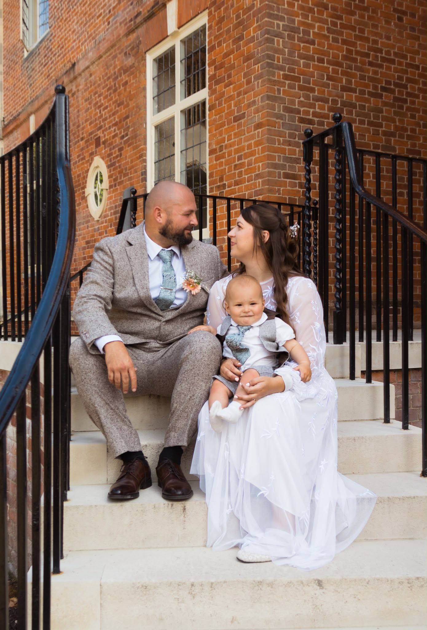Bride, groom and their baby sitting together on the stone steps of a wedding venue