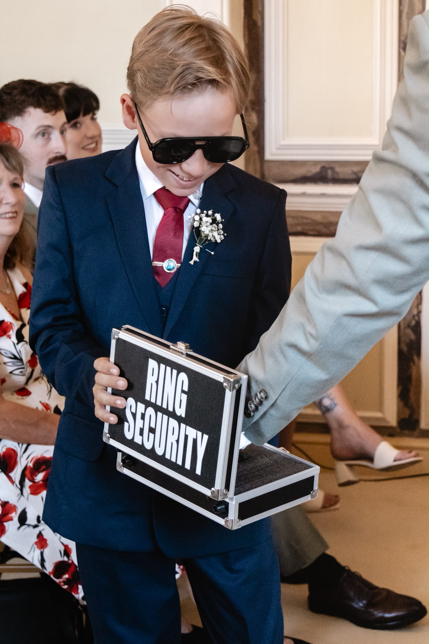 A young ring bearer in sunglasses carrying a Ring Security briefcase at a wedding