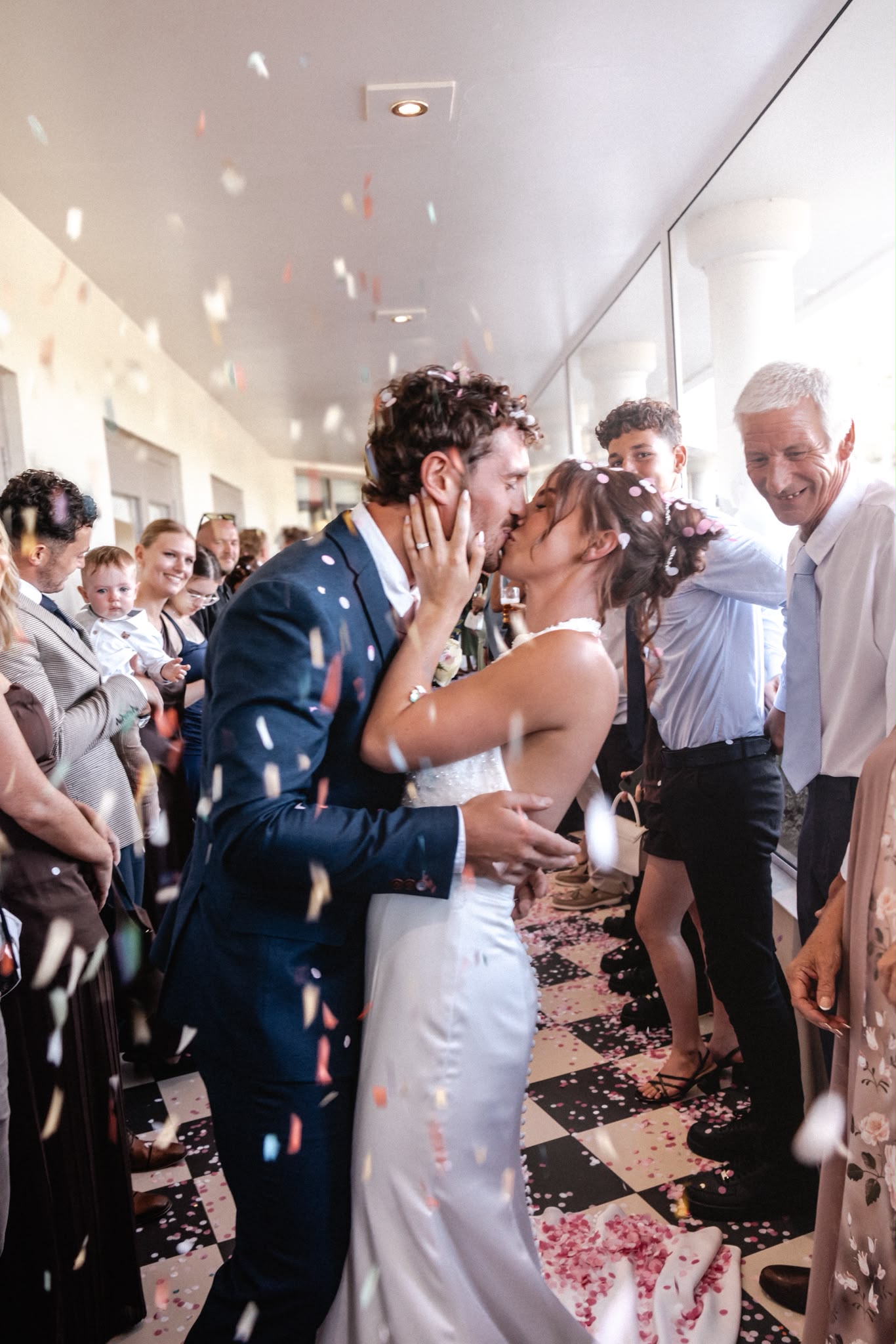Bride and groom kissing as confetti falls around them surrounded by wedding guests