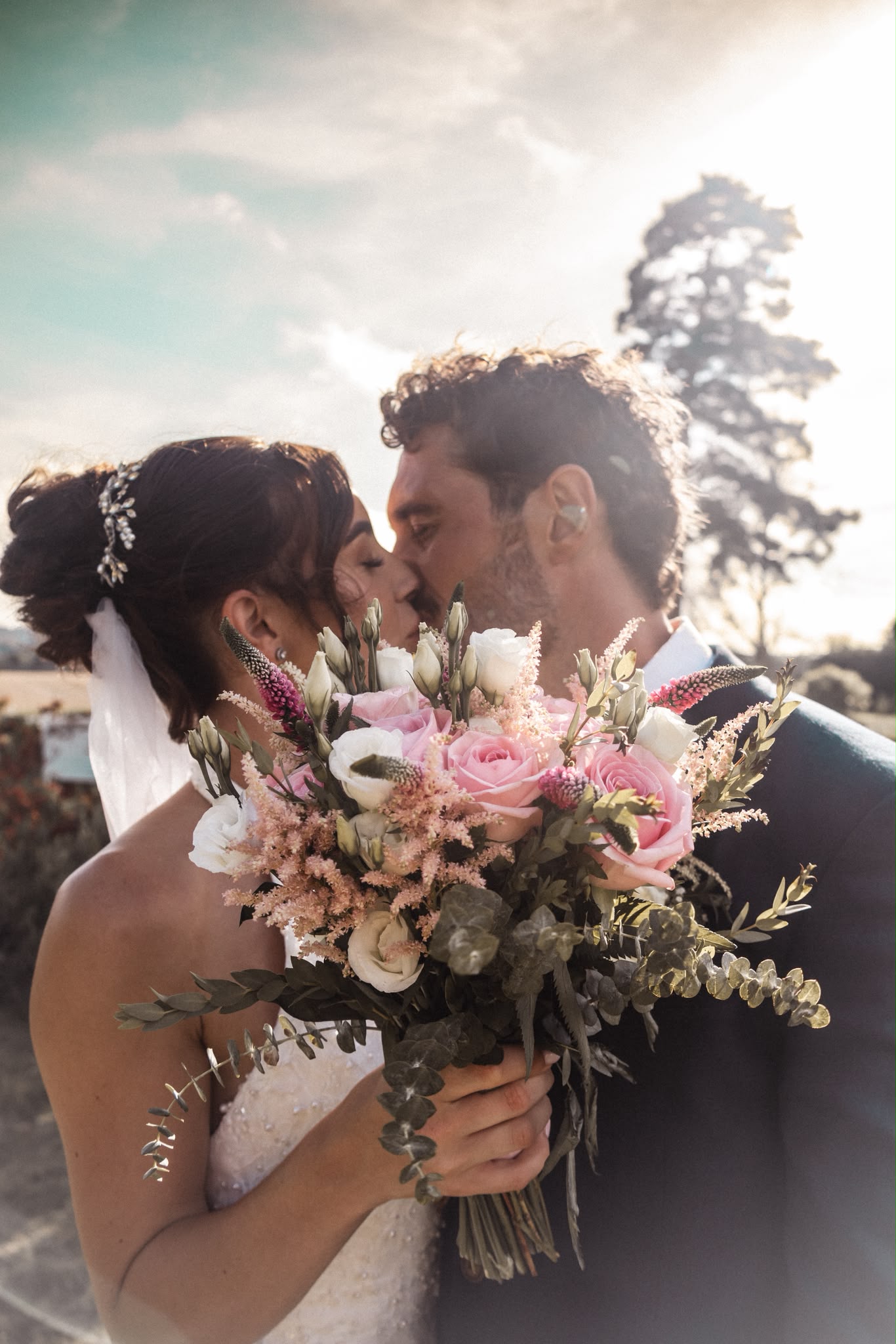 Bride and groom kissing in warm backlight, the bride holding a bouquet of pink and white roses and astilbe