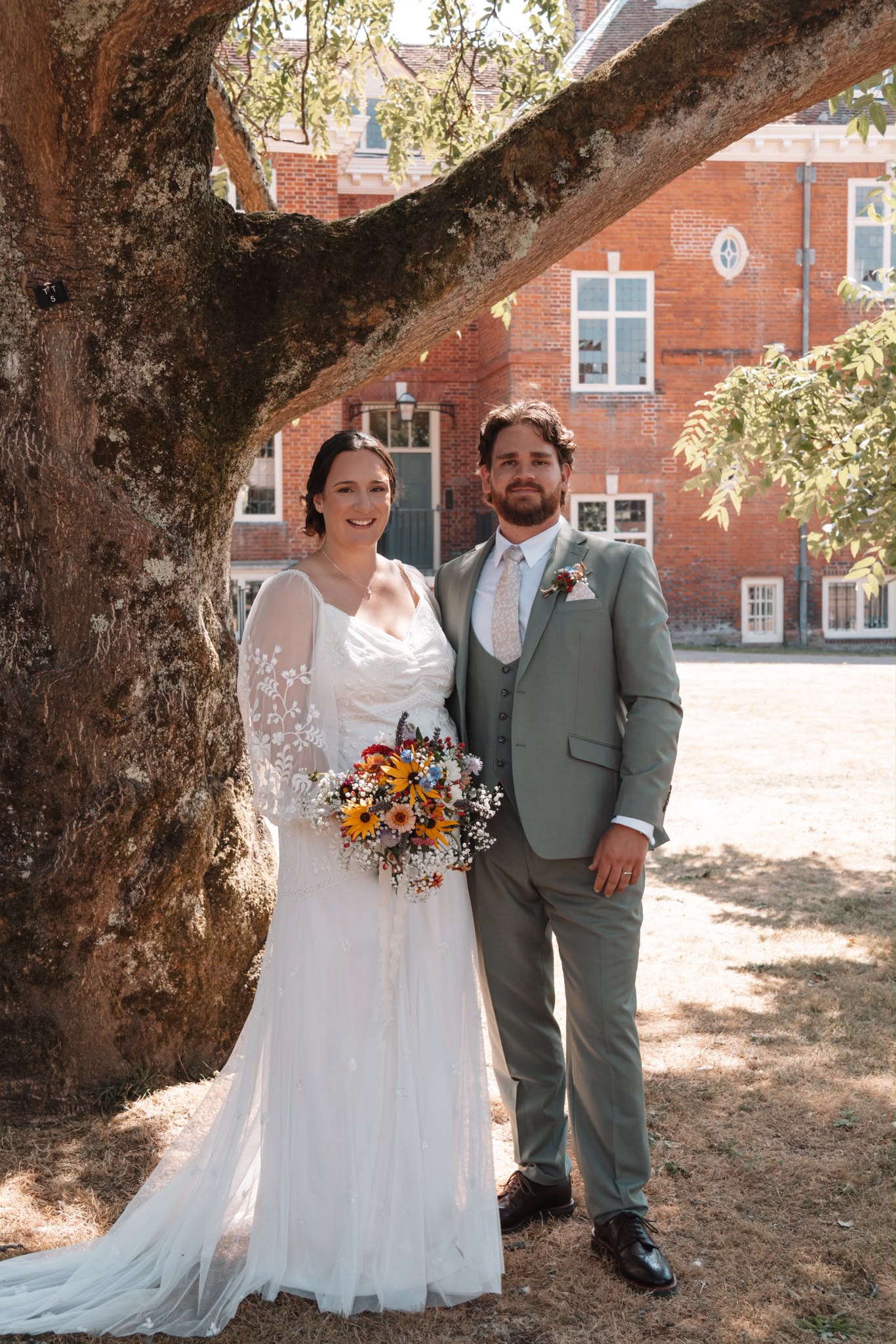 Bride and groom standing beneath a mature tree, the groom in a sage green three-piece suit