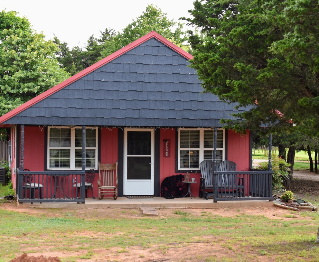 Prairie Willow cottage exterior with red siding, front porch and Adirondack chairs surrounded by trees