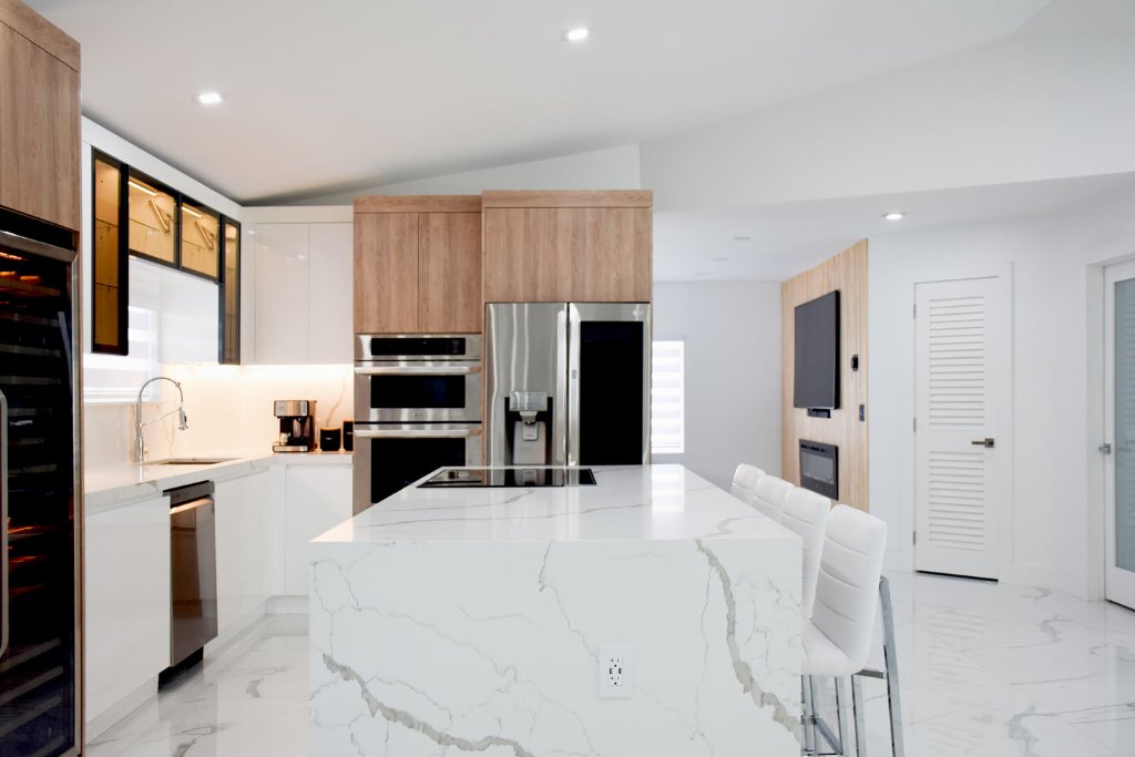 White Oak upper cabinets with high-gloss lacquered lowers and Calacatta quartz island — integrated stainless appliances and LED underlighting in a bright, open-plan kitchen.