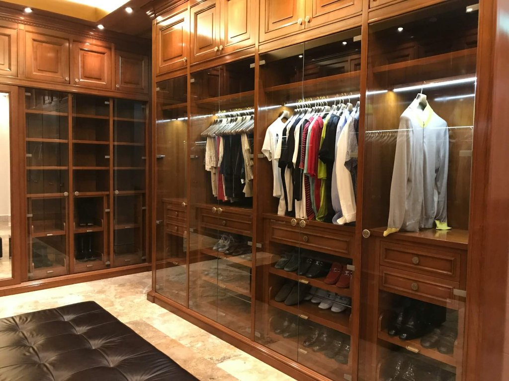 Solid walnut walk-in closet with glass-front cabinets, integrated LED lighting and brass hardware — your wardrobe, curated and lit like a boutique.