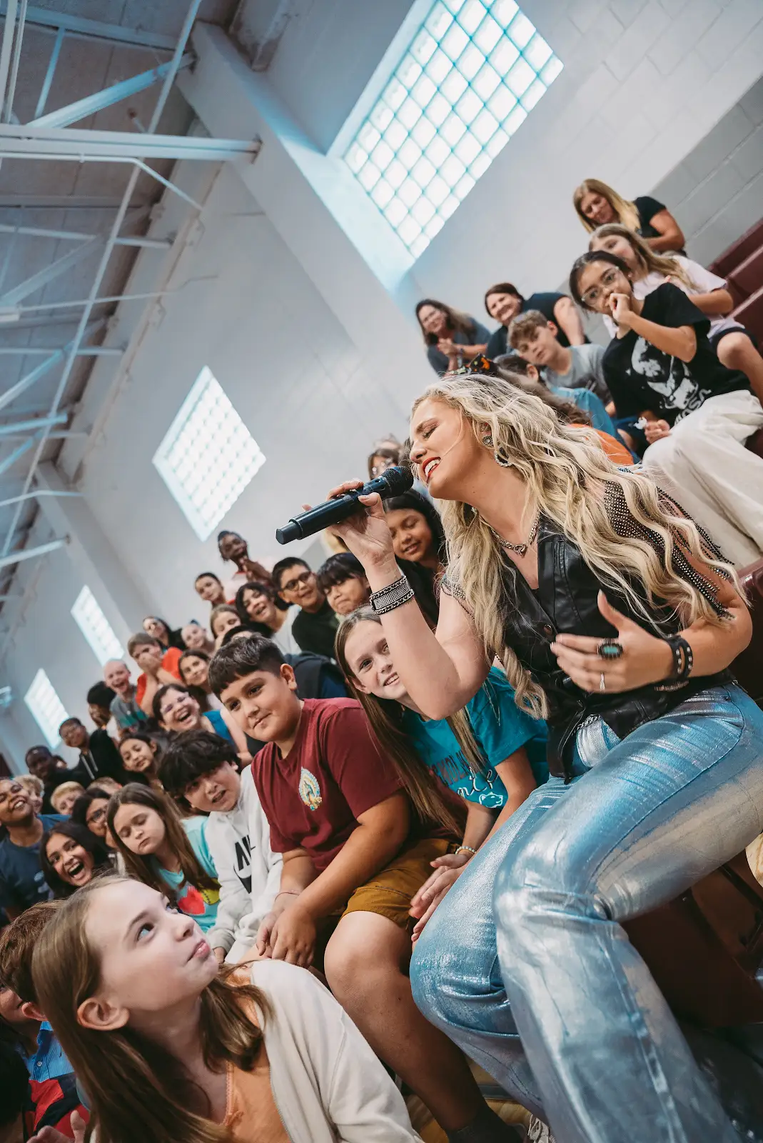 Kate Watson singing to students seated on gymnasium bleachers