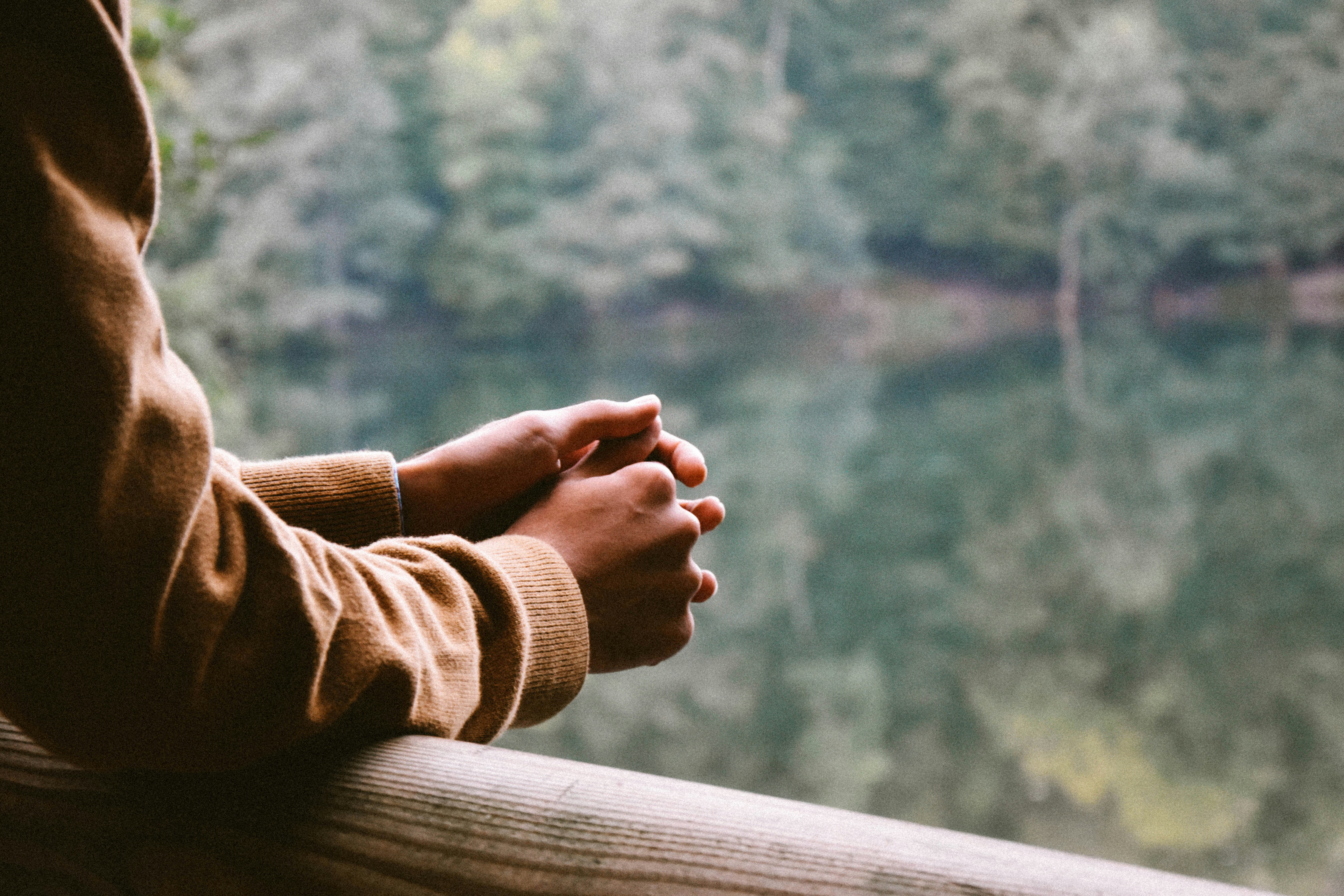 A person's clasped hands resting on a wooden railing, looking out over calm water and trees