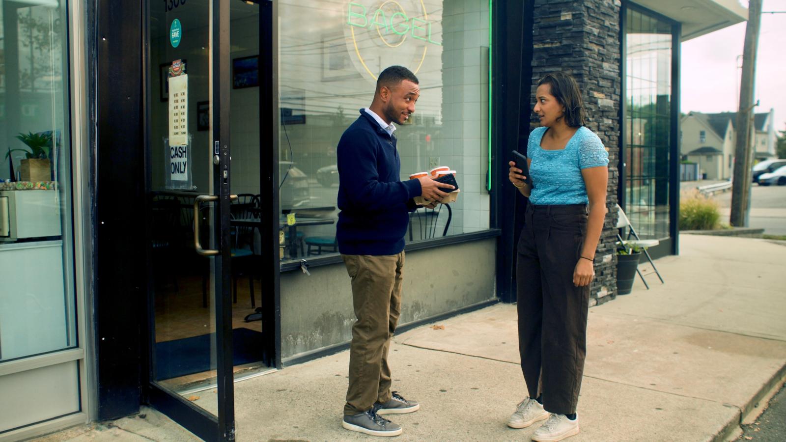 Two characters from the film Doctored standing on the sidewalk outside the More Than A Bagel storefront