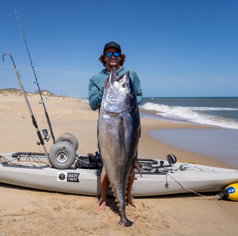Quentin Turko holding a large tuna on an Outer Banks beach with a Salty Crew kayak